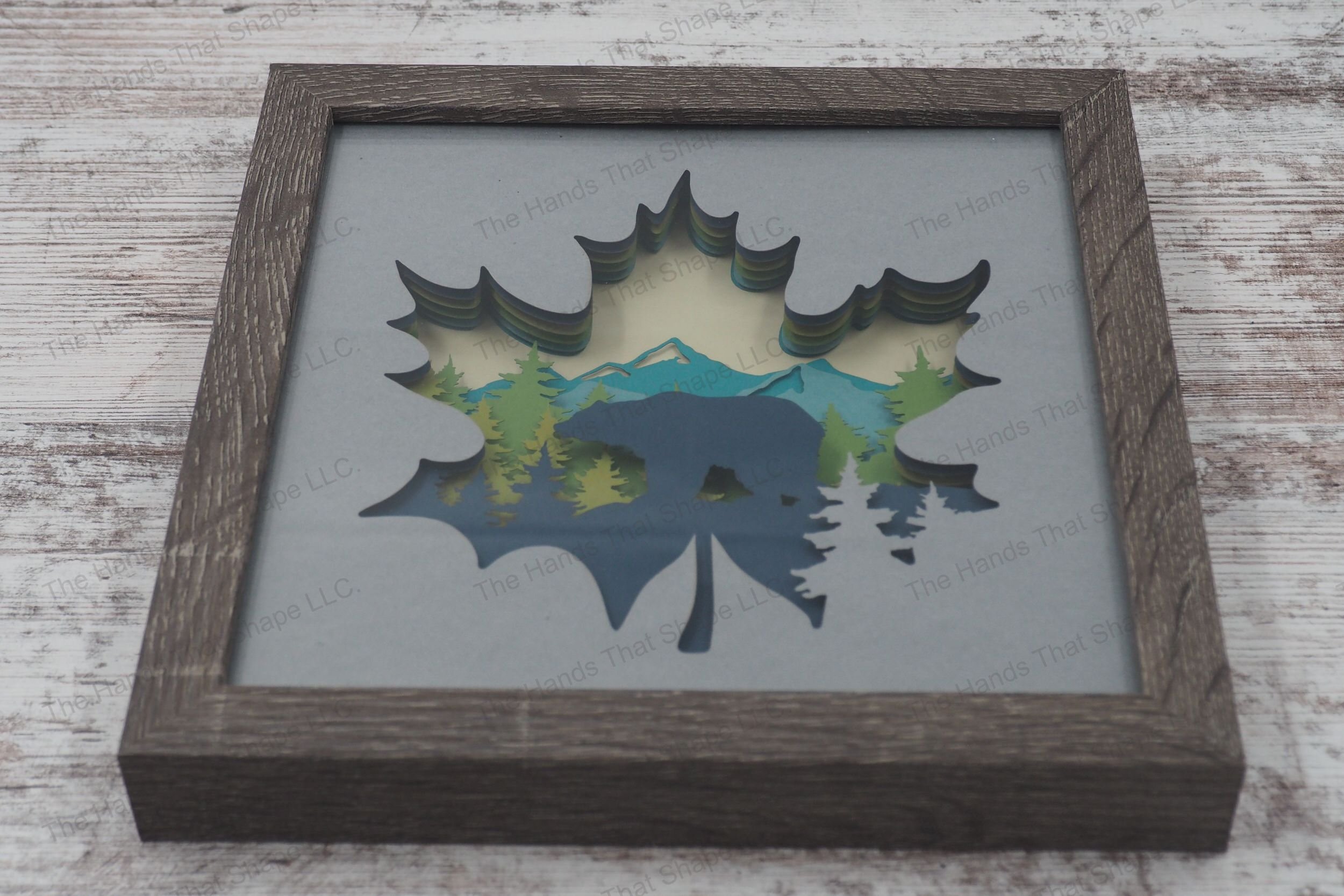 A shadowbox depicting a bear silhouette amidst pine trees and mountains, framed within a maple leaf, crafted from layers of cardstock in a gray MDF frame.