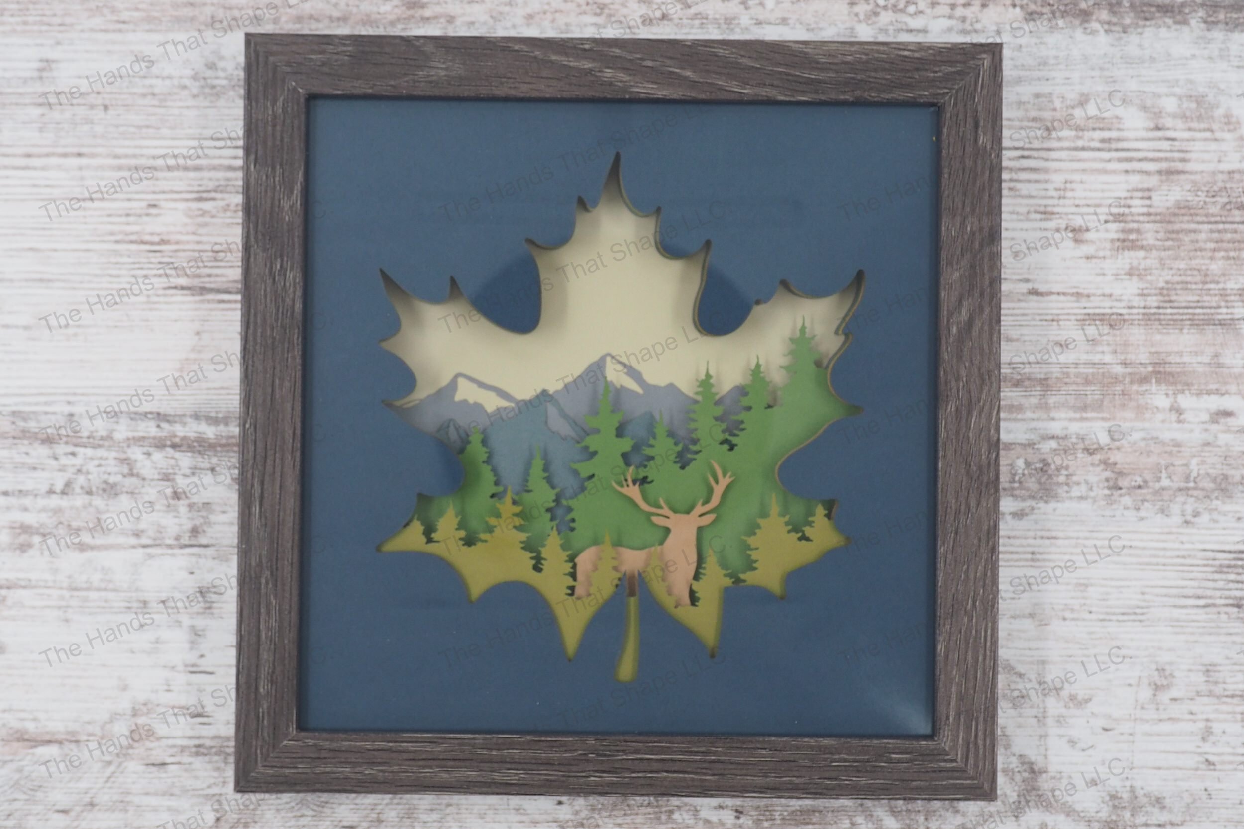 An layered paper art piece showcasing a buck silhouette with pine trees and a mountain background, all contained within a maple leaf shape, in a gray MDF frame.