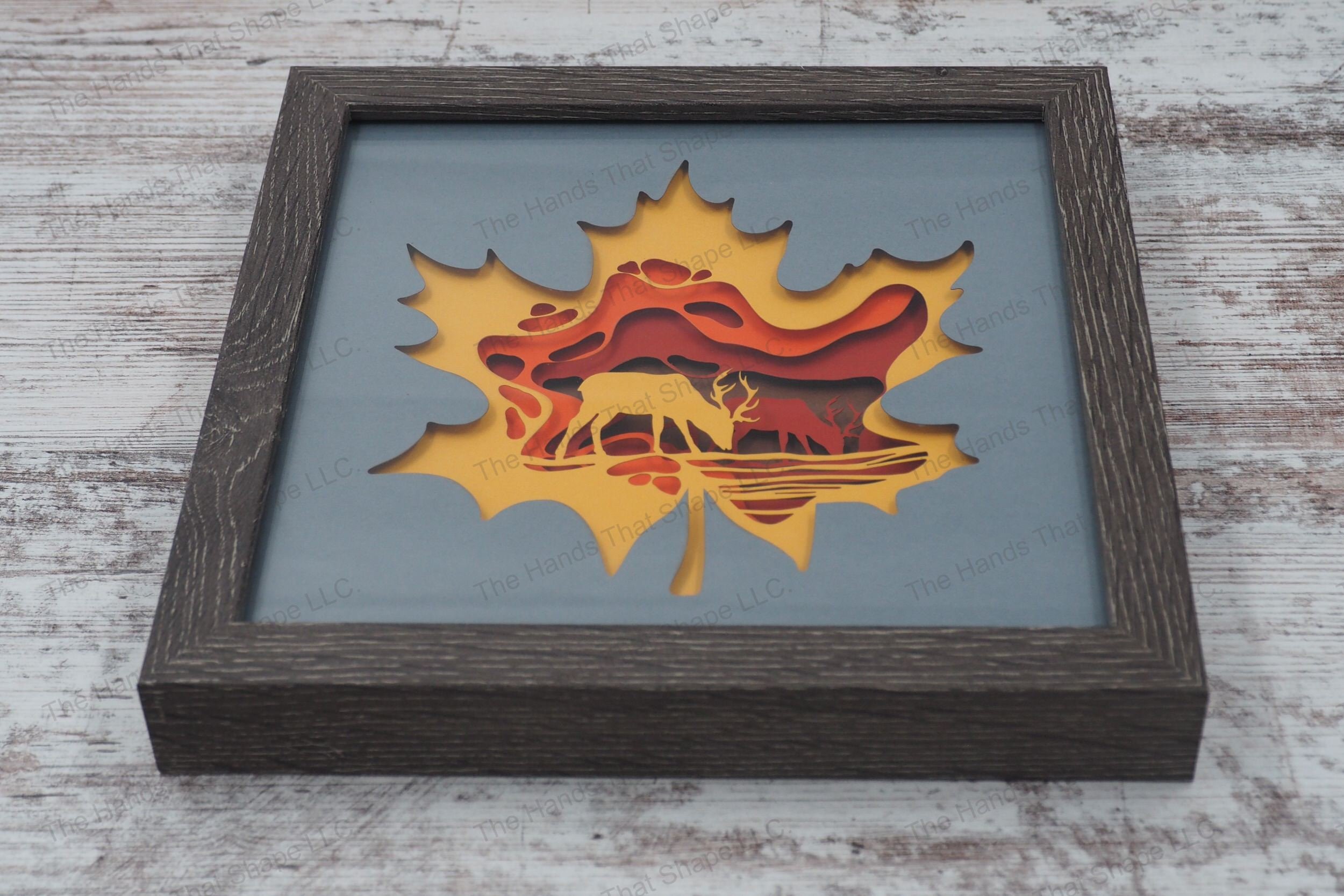 A shadowbox featuring two bucks by a stream surrounded by fall colors, encapsulated in a maple leaf outline, assembled from cardstock in a gray frame.