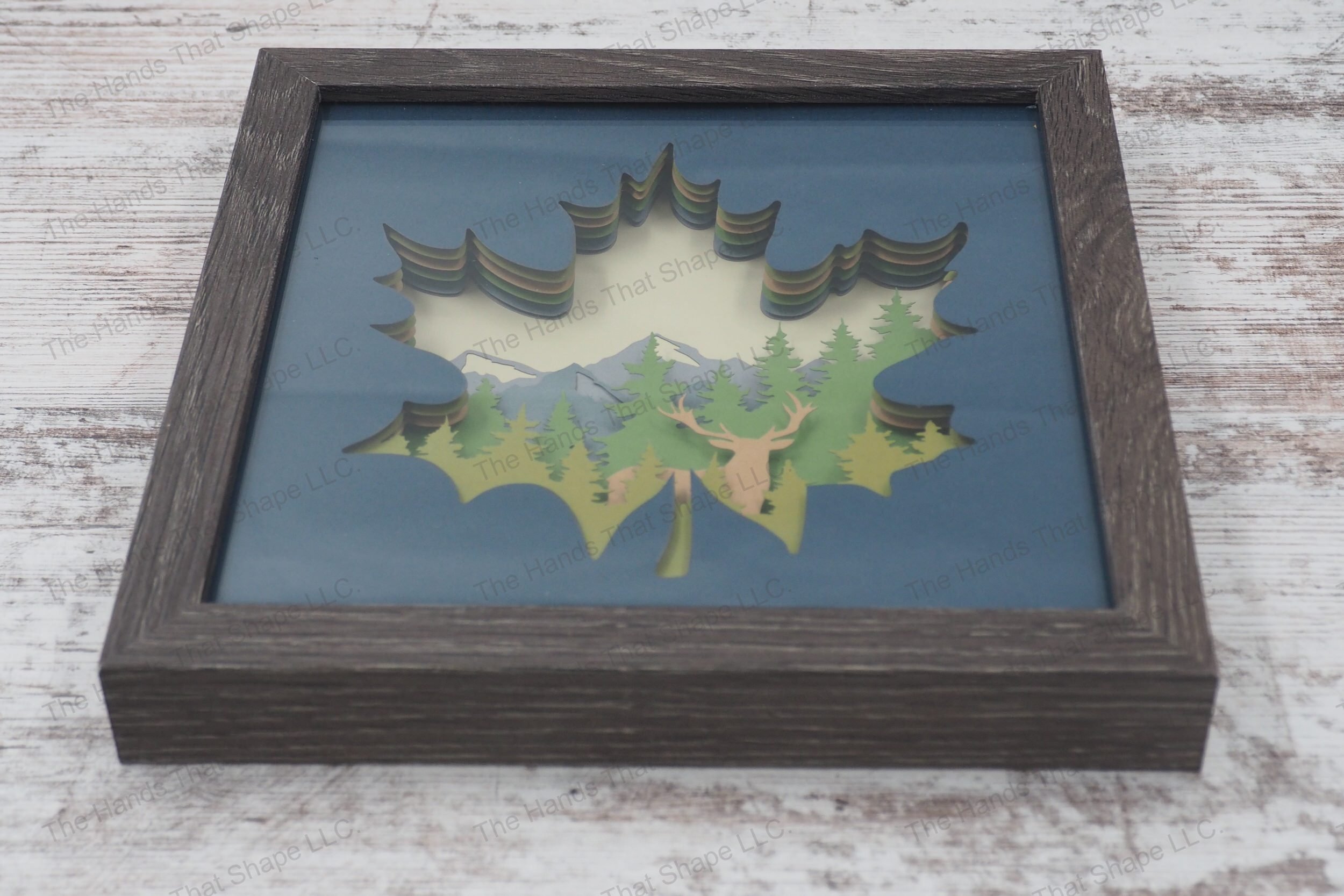 An layered paper art piece showcasing a buck silhouette with pine trees and a mountain background, all contained within a maple leaf shape, in a gray MDF frame.