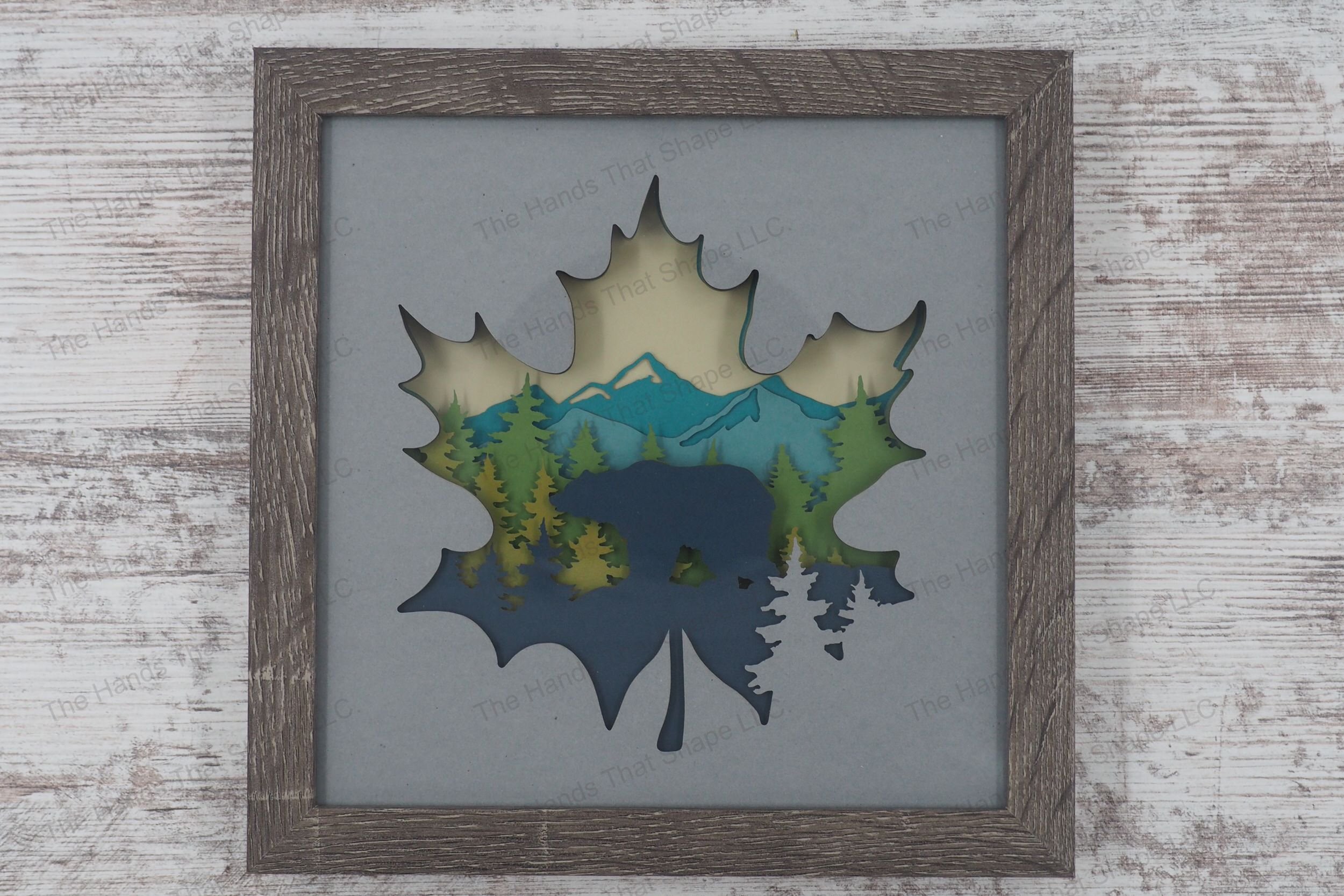 A shadowbox depicting a bear silhouette amidst pine trees and mountains, framed within a maple leaf, crafted from layers of cardstock in a gray MDF frame.
