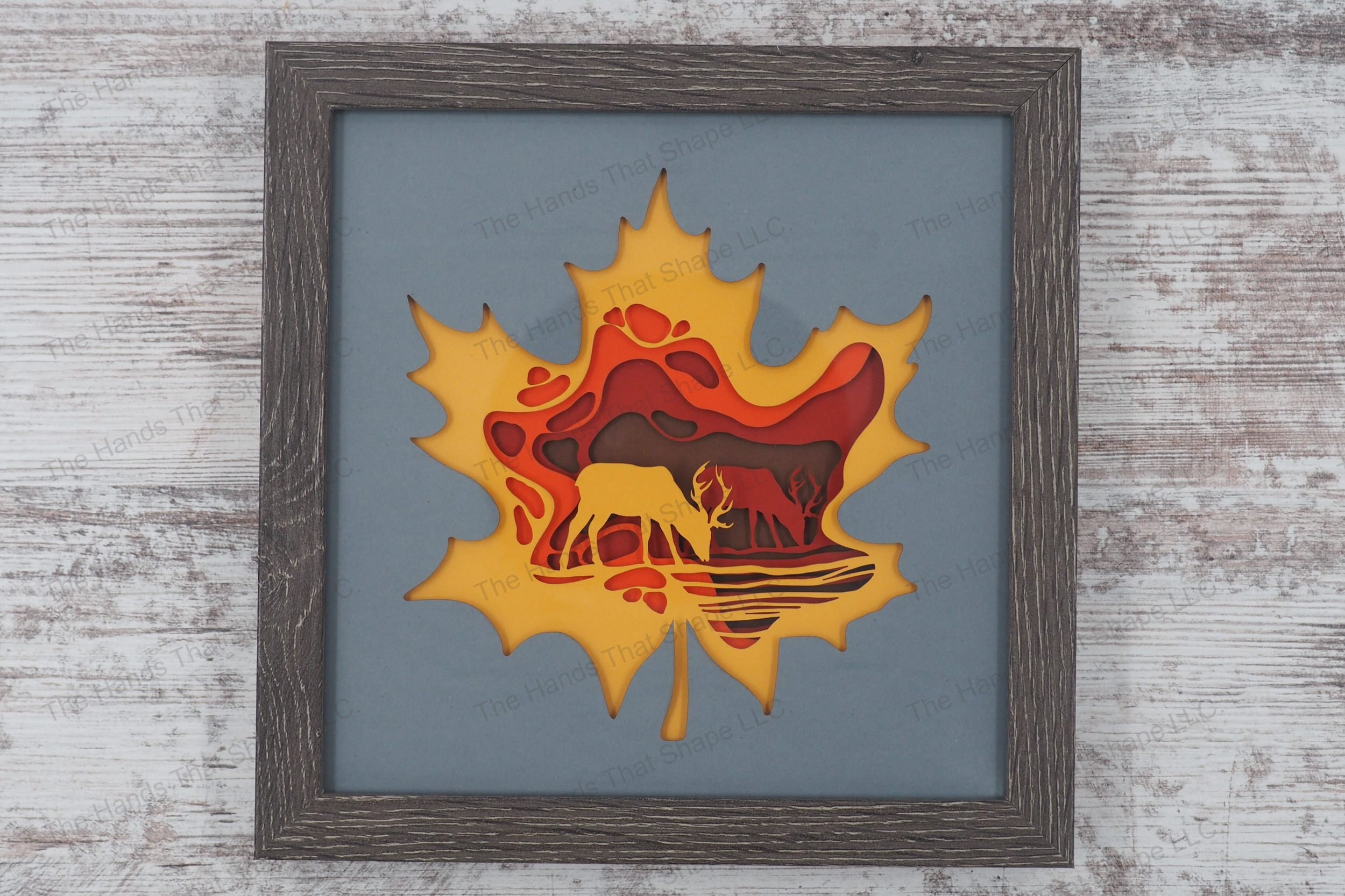 A shadowbox featuring two bucks by a stream surrounded by fall colors, encapsulated in a maple leaf outline, assembled from cardstock in a gray frame.