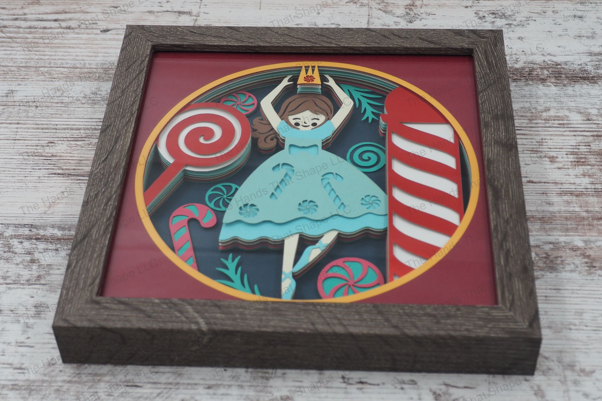 A whimsical framed layered paper shadowbox depicting a Nutcracker-inspired ballerina with a candy-themed dress, surrounded by pinwheels and candy canes.