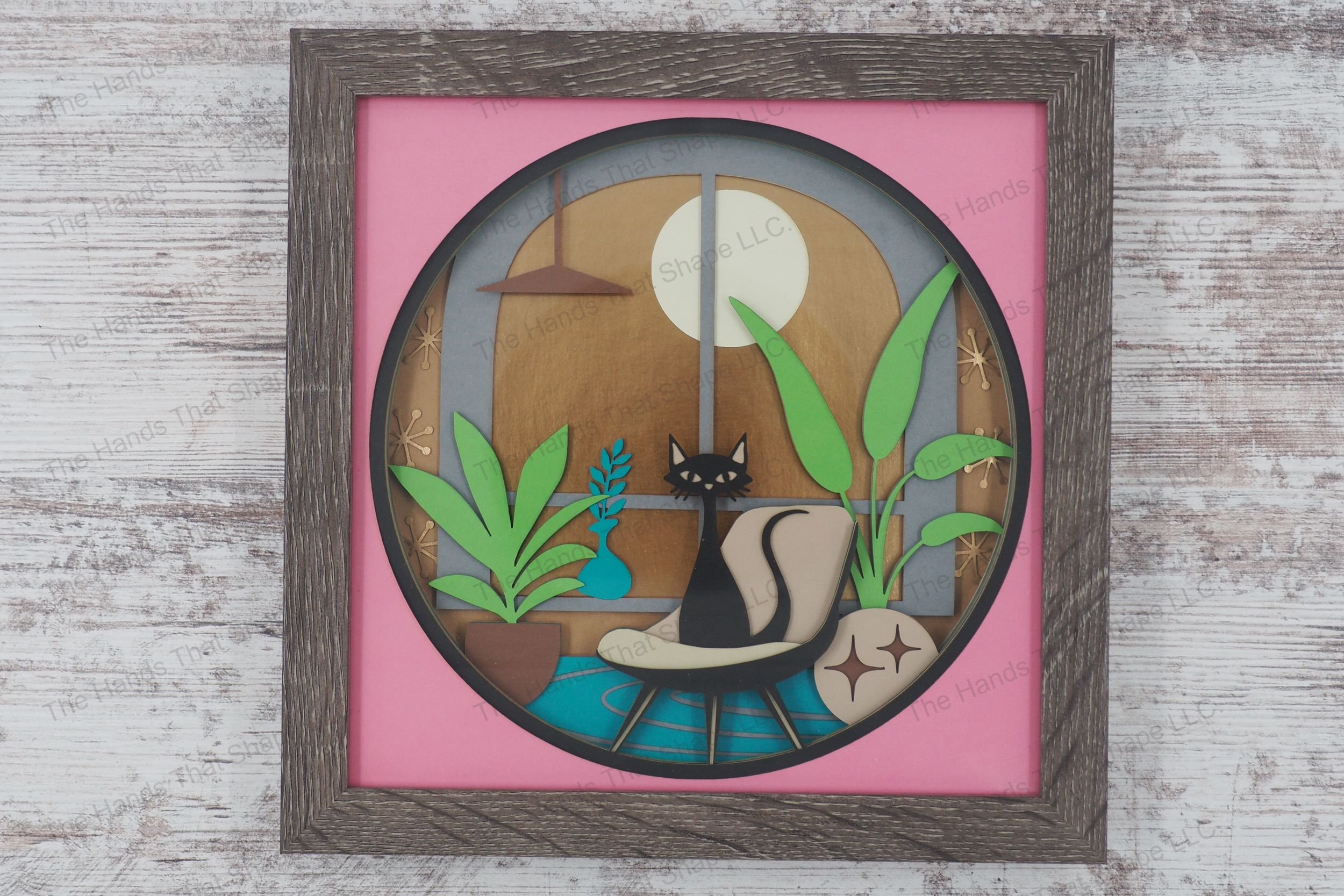 A serene framed layered paper shadowbox depicting a cat sitting on a chair in a bohemian living room with potted plants and a large window showing a golden sky.