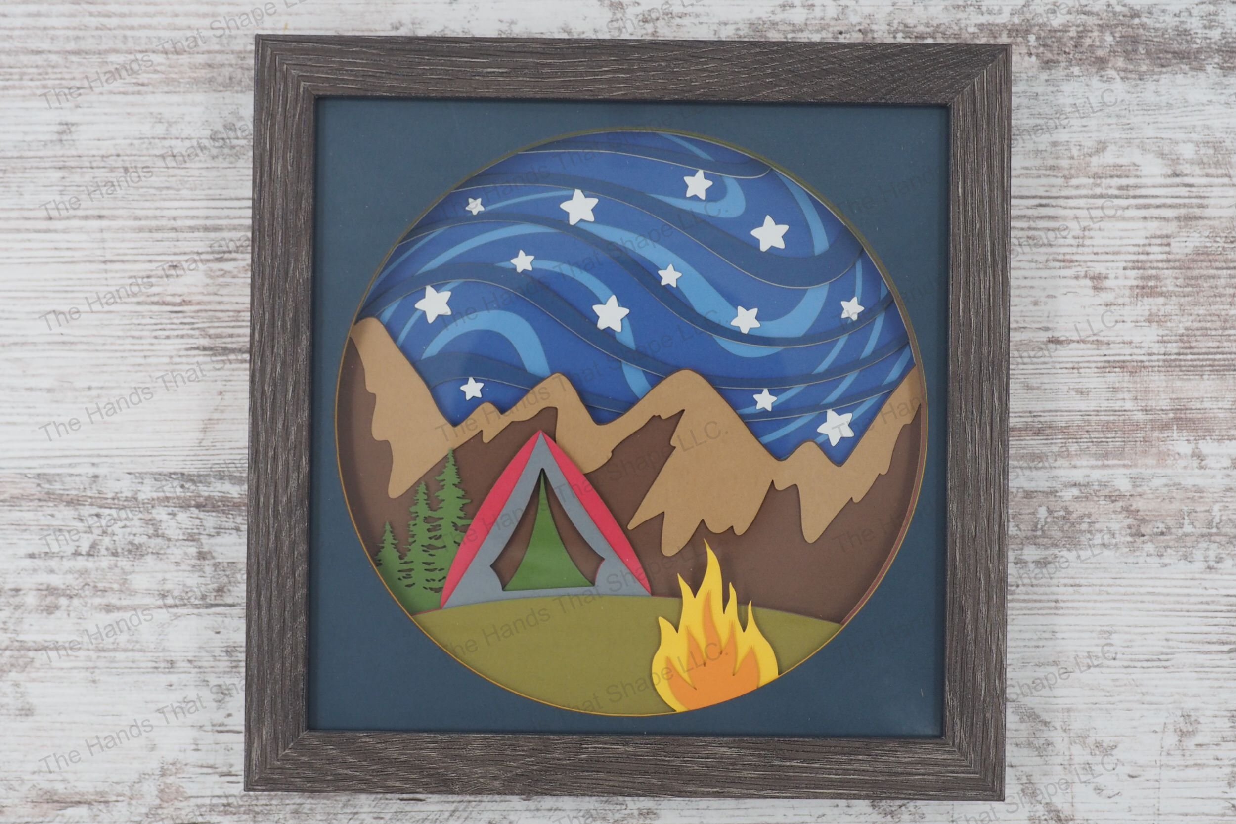 A framed layered paper shadowbox depicting a camping scene with a tent, campfire, and pine trees under a starry night sky and mountain range.