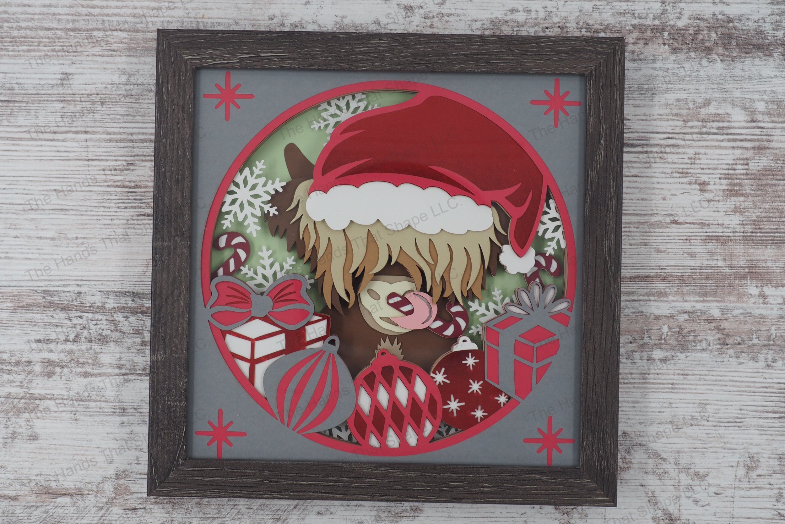 A festive framed shadowbox depicting a baby Highland cow in a Christmas hat, with presents, ornaments, and candy canes, crafted from 14 layers of cardstock.