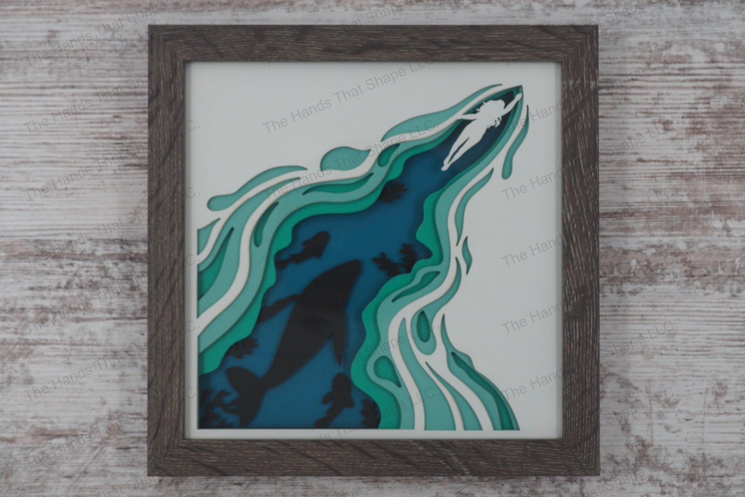 A framed layered paper shadowbox depicting a woman swimming through the ocean, revealing silhouettes of marine life below, crafted from 5 layers of cardstock.
