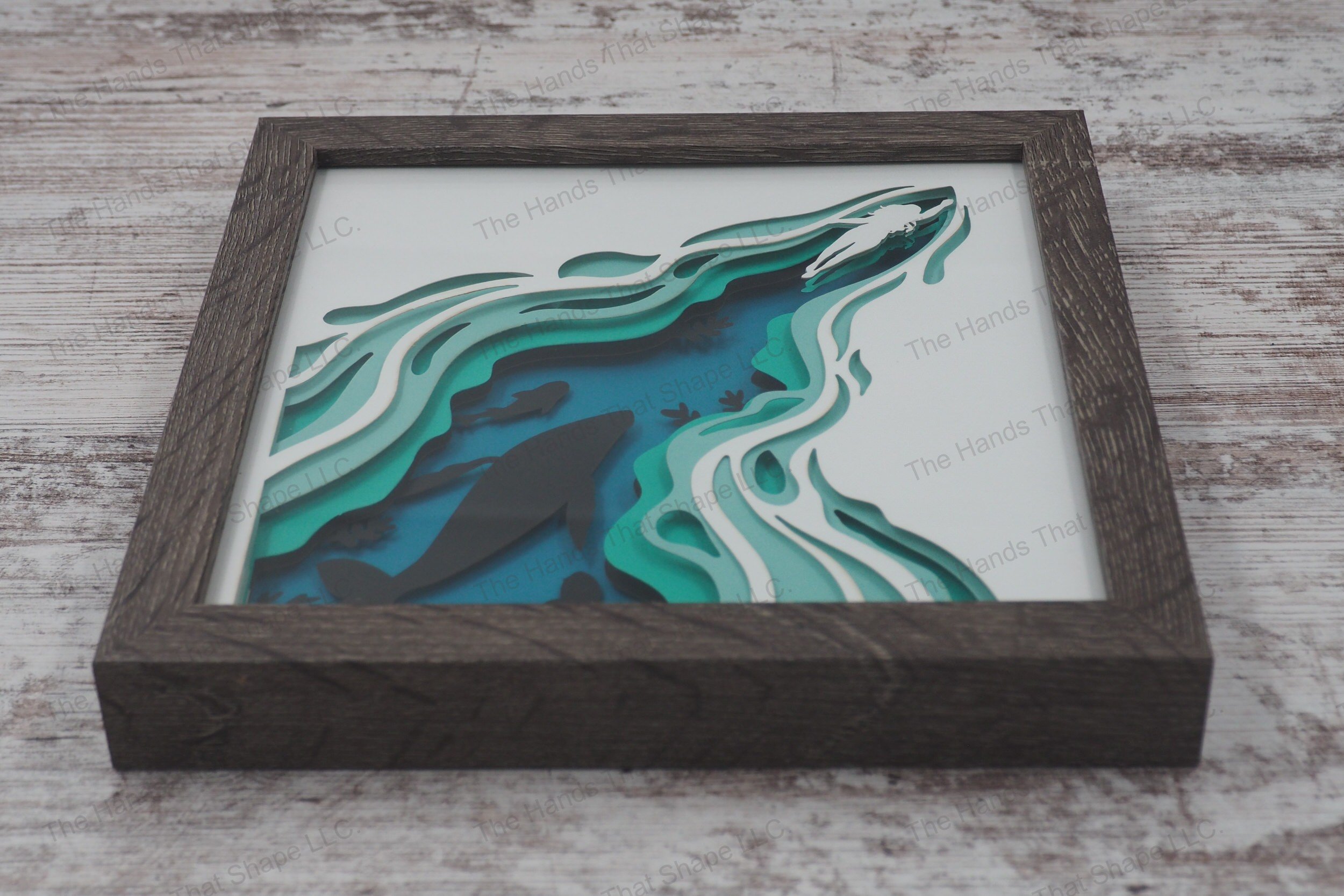 A framed layered paper shadowbox depicting a woman swimming through the ocean, revealing silhouettes of marine life below, crafted from 5 layers of cardstock.
