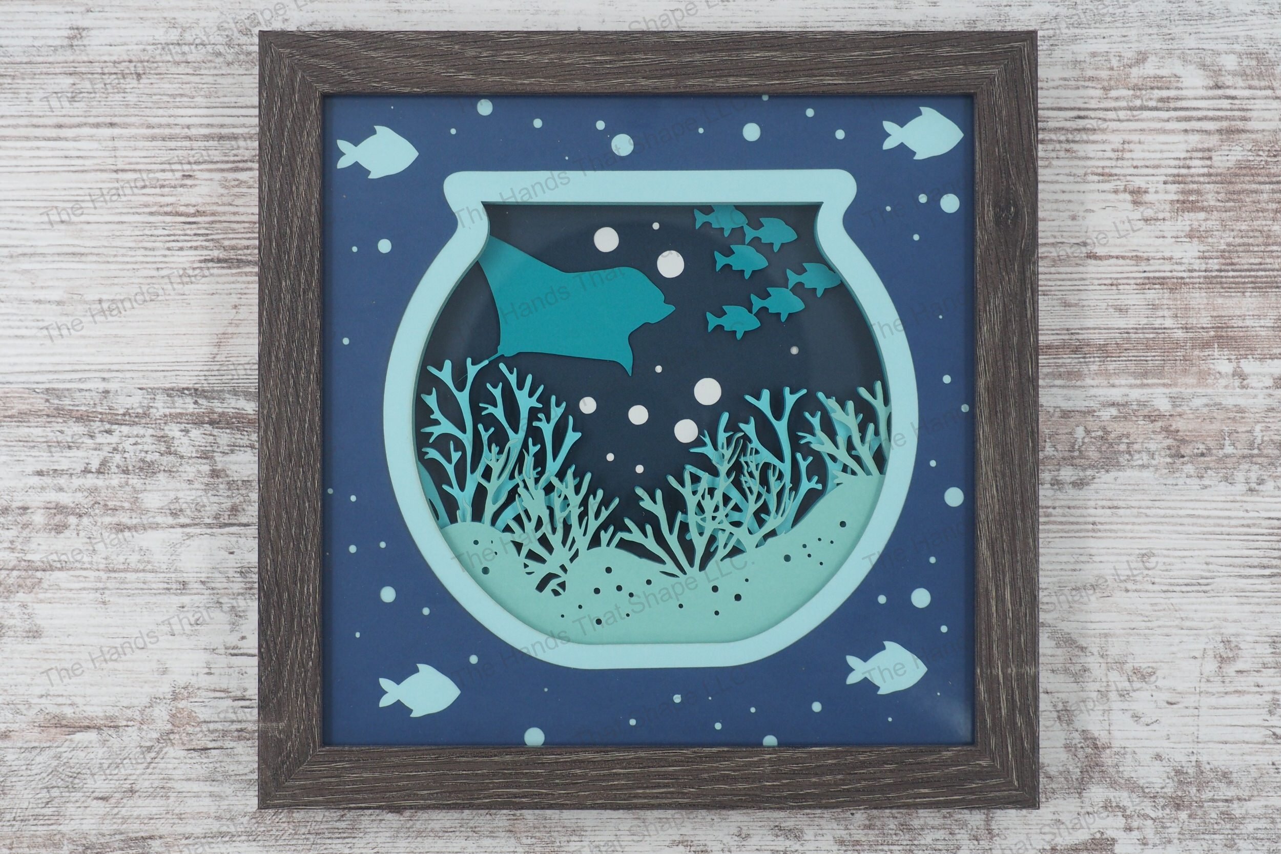 A framed layered paper shadowbox depicting an underwater ocean scene within a fishbowl shape, featuring coral, a stingray, school of fish, and bubbles.