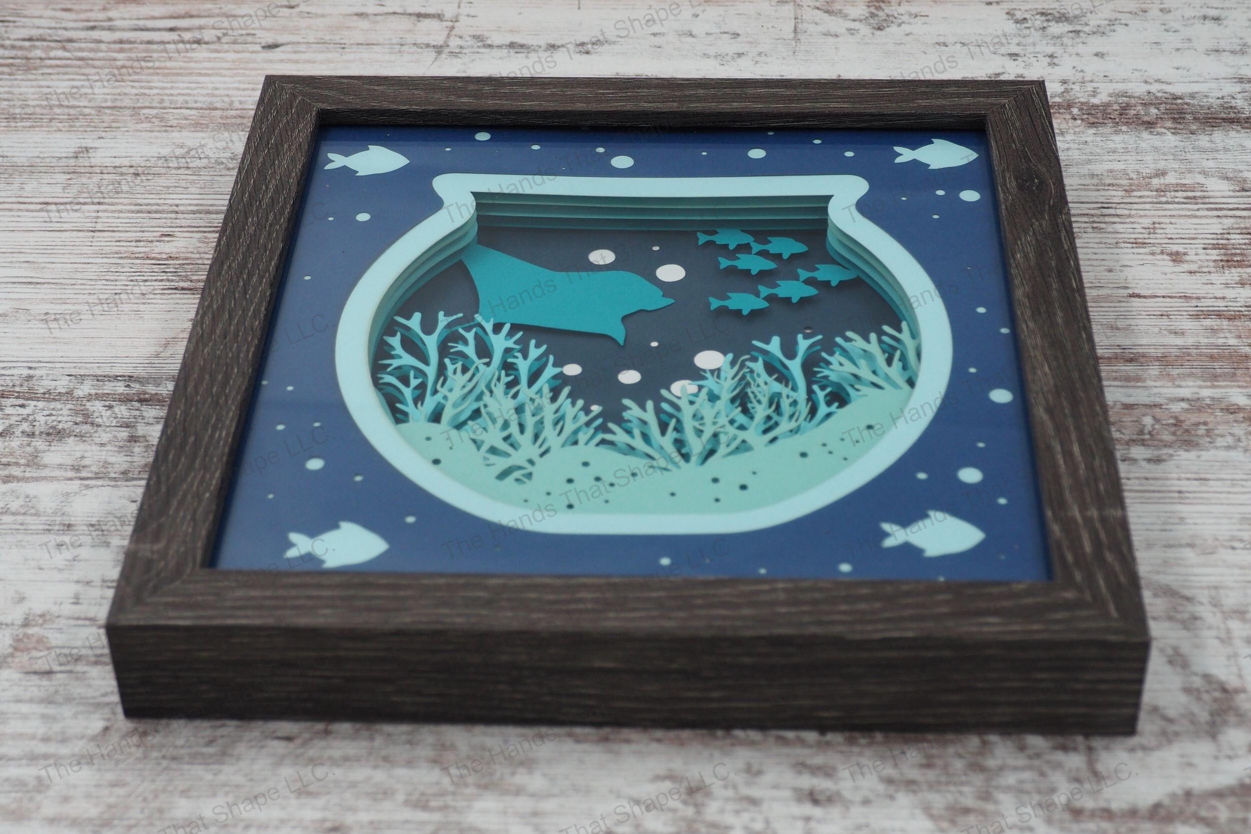 A framed layered paper shadowbox depicting an underwater ocean scene within a fishbowl shape, featuring coral, a stingray, school of fish, and bubbles.