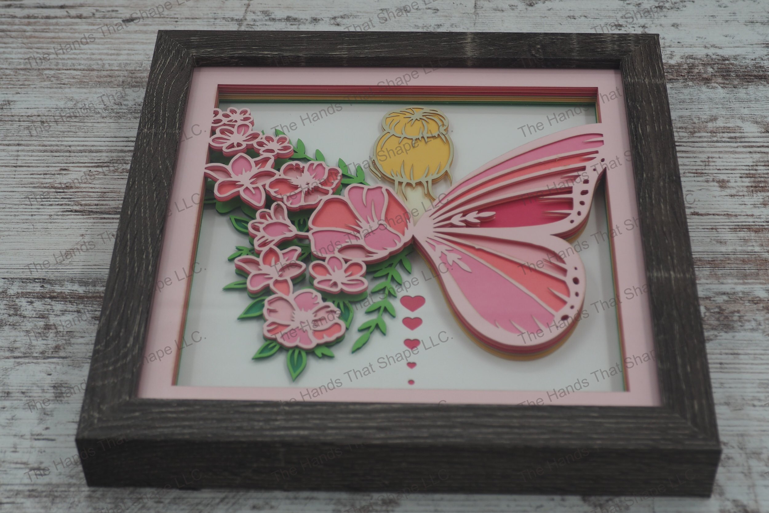 A layered paper shadowbox depicting a forest fairy with one butterfly wing and another made of flowers, crafted from 12 layers of cardstock in a gray MDF frame.