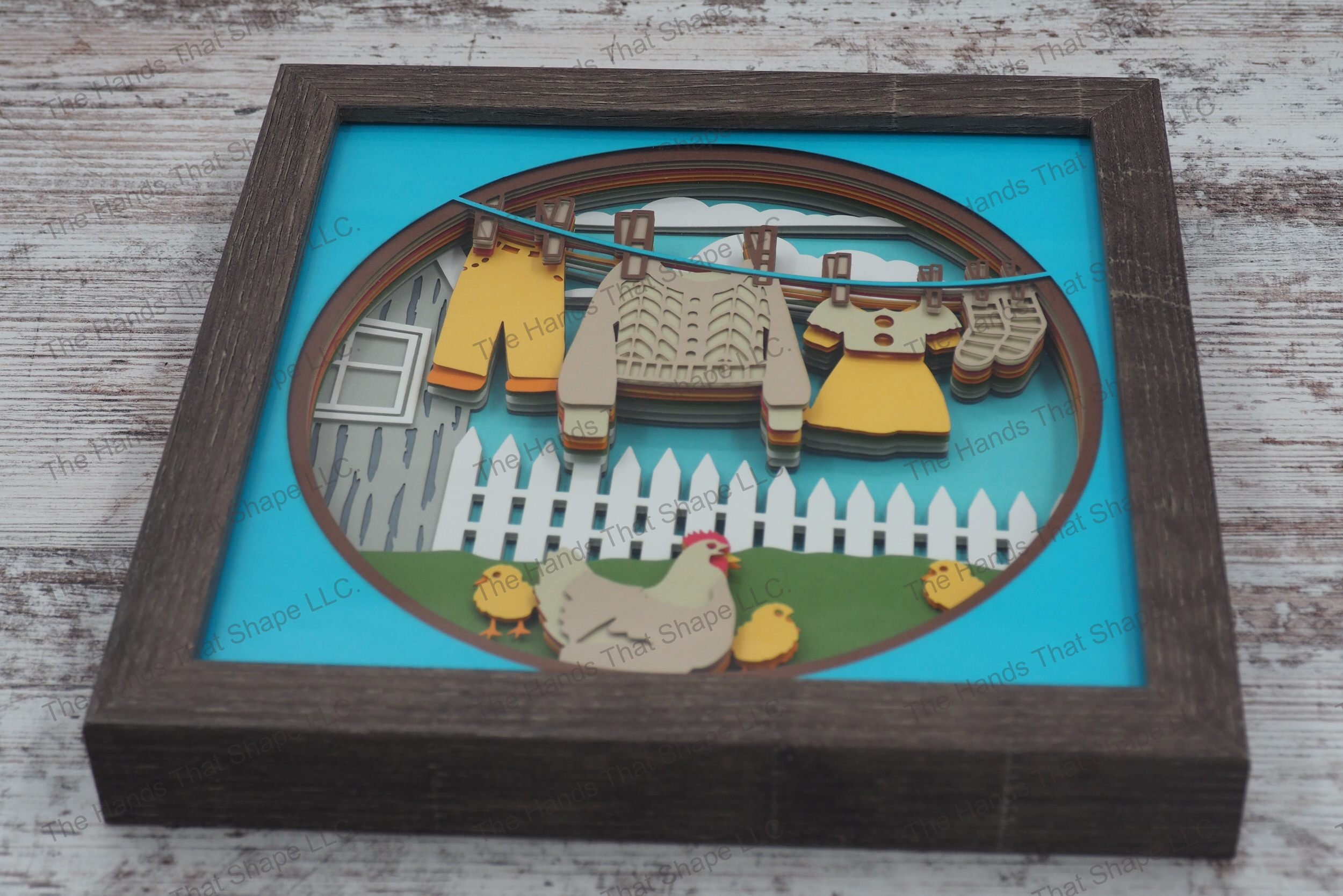 A framed layered paper shadowbox depicting a charming farmhouse scene with clothes drying on a line, a chicken with chicks, and a white picket fence.