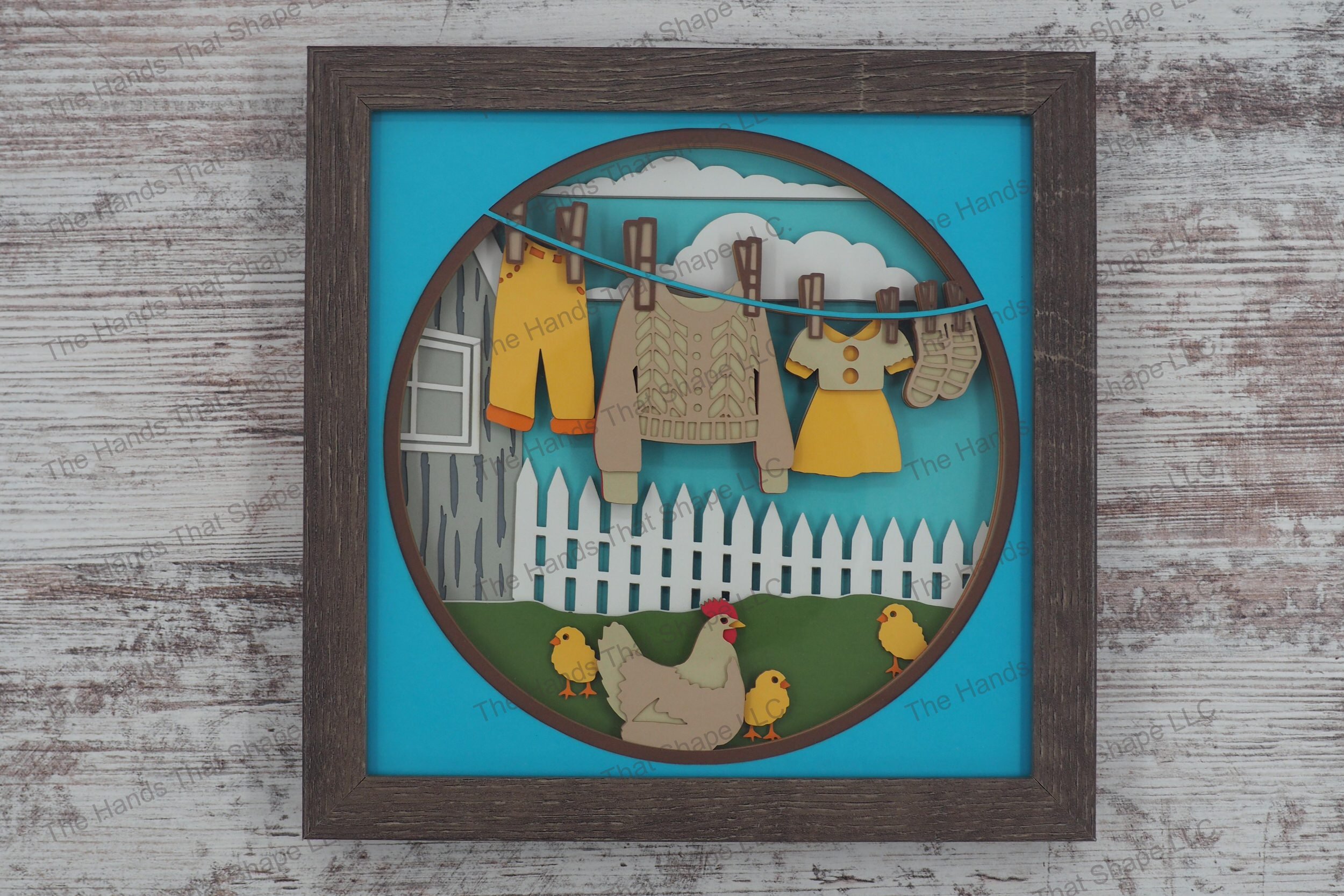 A framed layered paper shadowbox depicting a charming farmhouse scene with clothes drying on a line, a chicken with chicks, and a white picket fence.