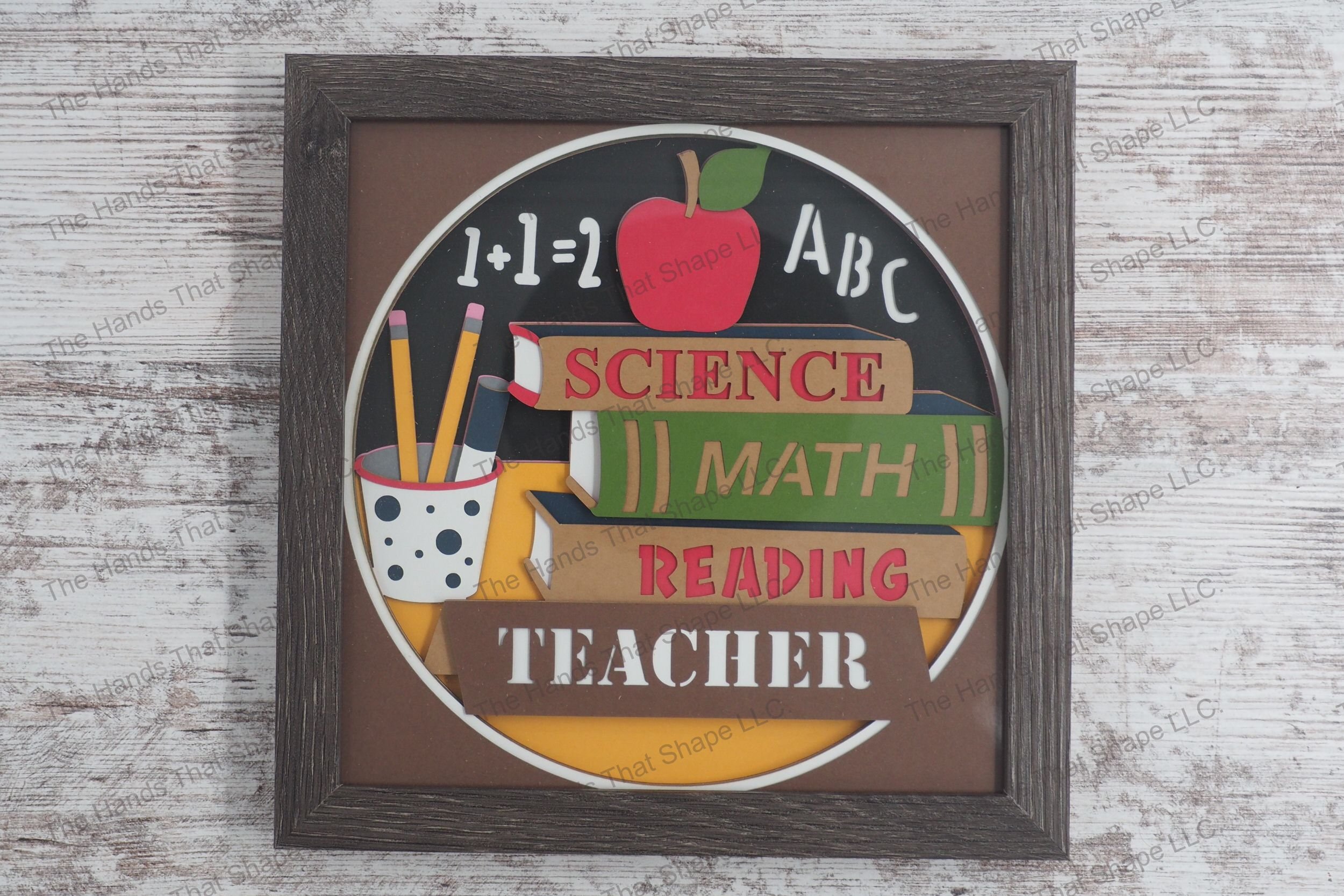 A framed layered paper shadowbox depicting a teacher's desk scene with books, an apple, pencils, and a chalkboard featuring basic math and literacy.