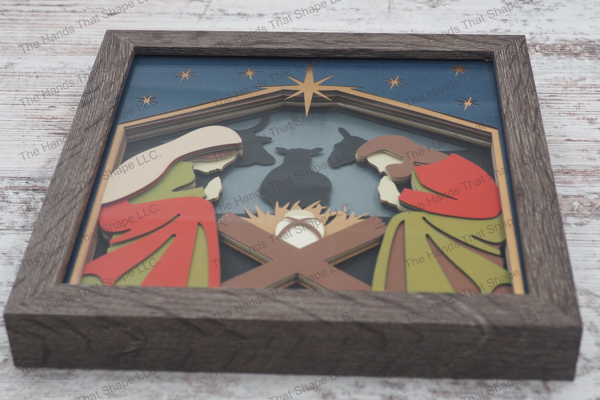 A framed layered paper shadowbox depicting the nativity scene with Mary, Joseph, and the baby Jesus, enhanced by animal silhouettes and a bright star.