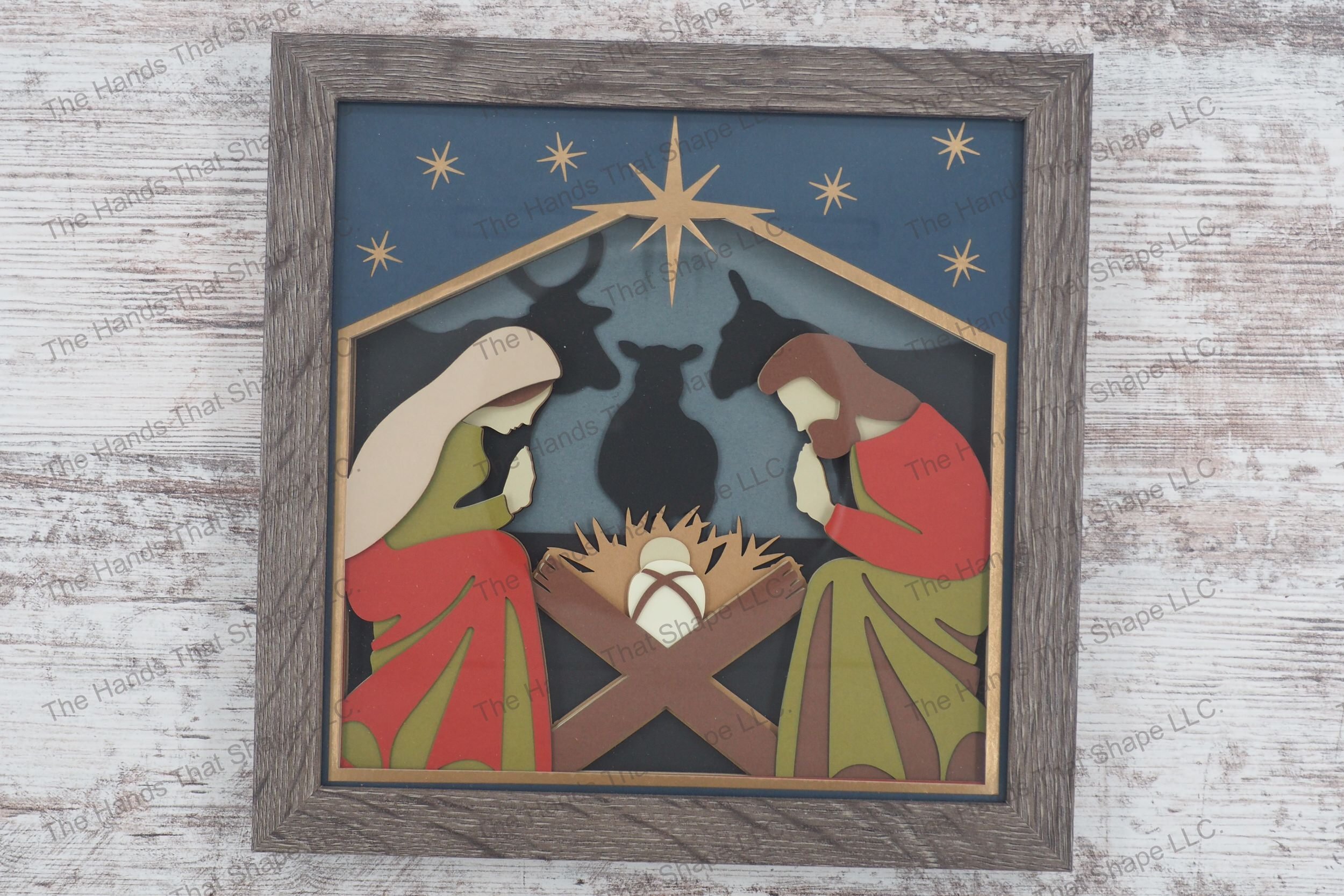 A framed layered paper shadowbox depicting the nativity scene with Mary, Joseph, and the baby Jesus, enhanced by animal silhouettes and a bright star.