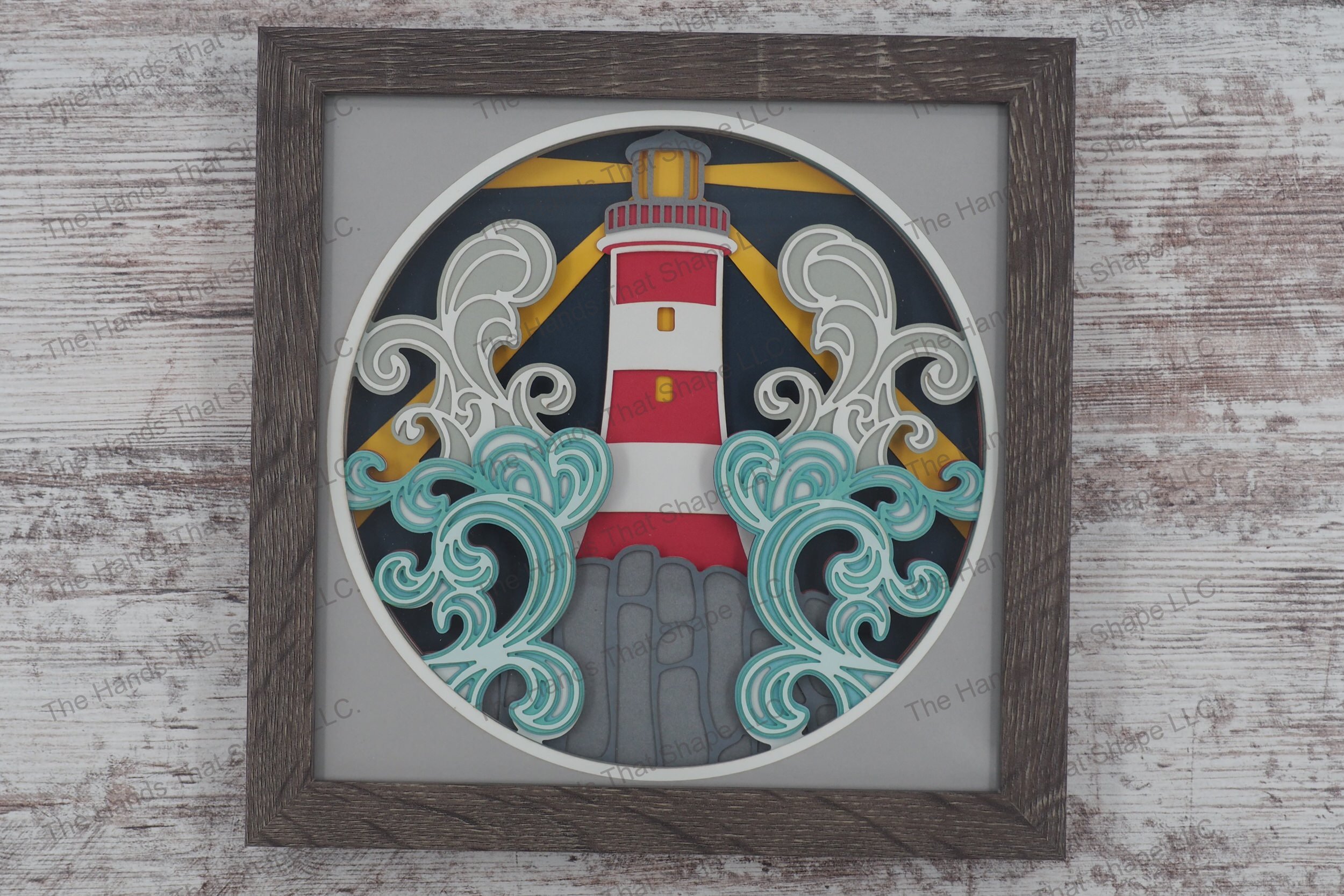 A framed layered paper shadowbox depicting a lighthouse on a rocky outcrop with crashing waves and light rays cutting through the night, crafted from cardstock.