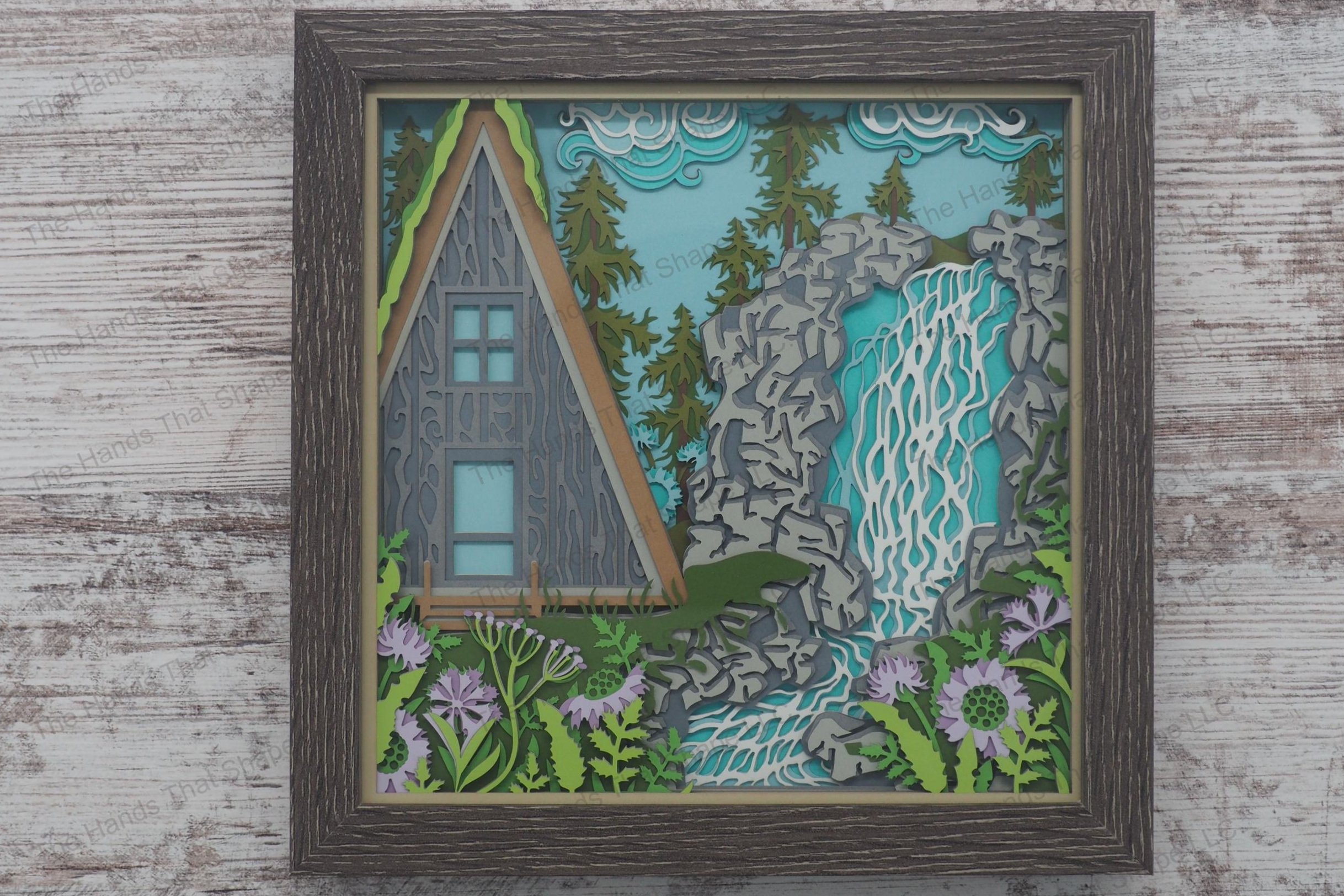 A framed layered paper shadowbox depicting an A-frame cabin by a waterfall and stream, surrounded by flower gardens and a pine forest.
