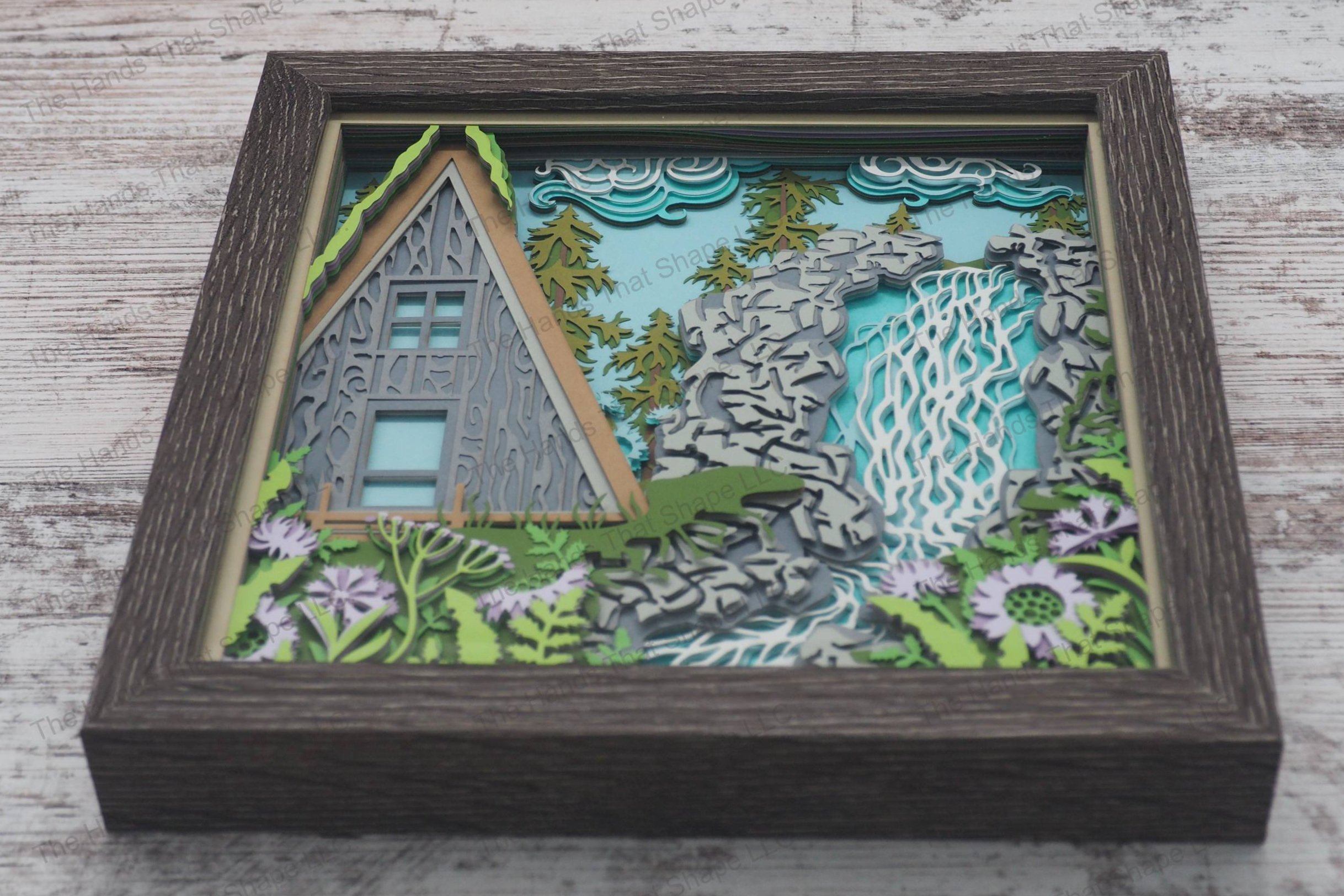 A framed layered paper shadowbox depicting an A-frame cabin by a waterfall and stream, surrounded by flower gardens and a pine forest.