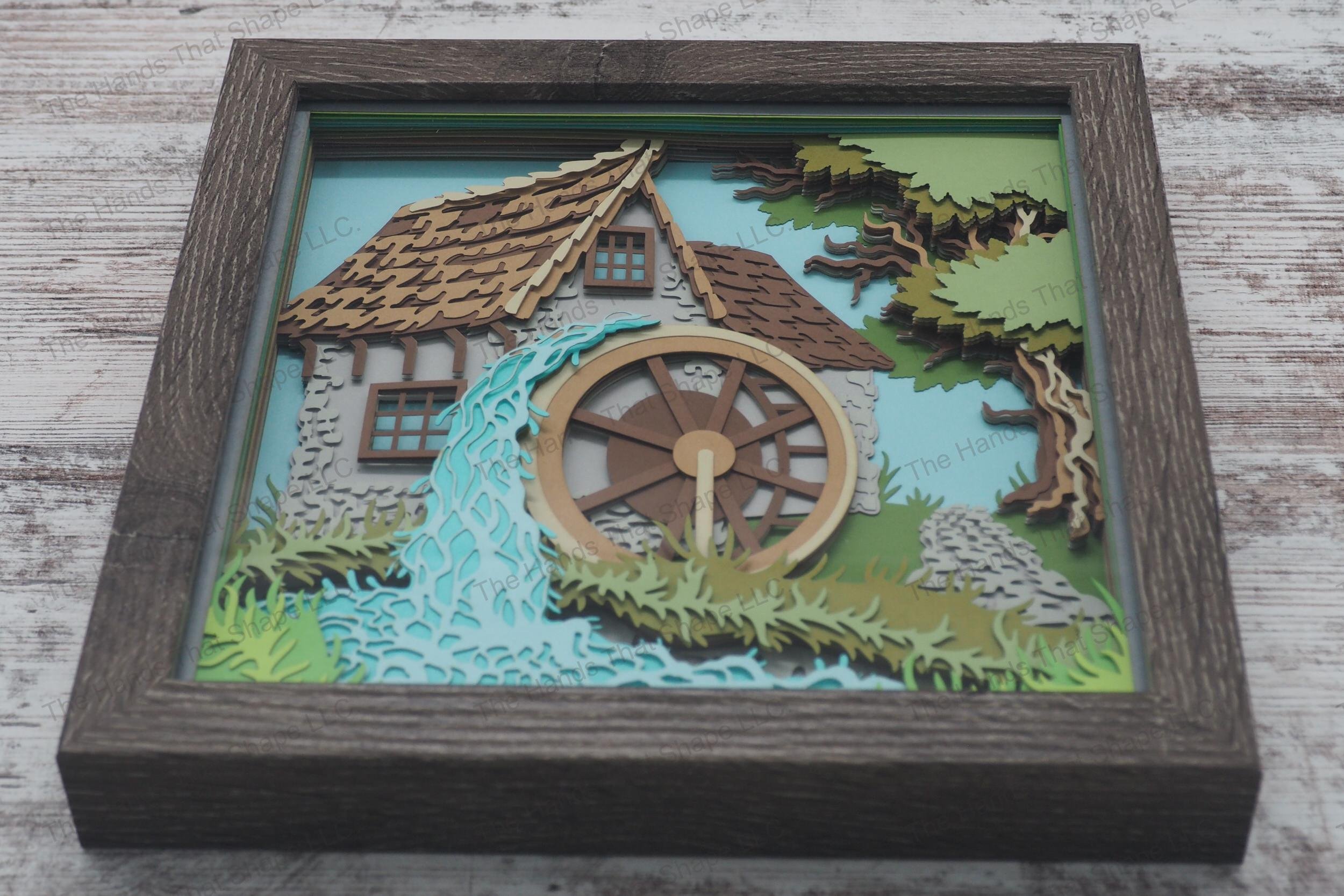 A framed layered paper shadowbox depicting a rustic water mill with a shake roof, stone walls, and water flowing over the wheel by a stream.