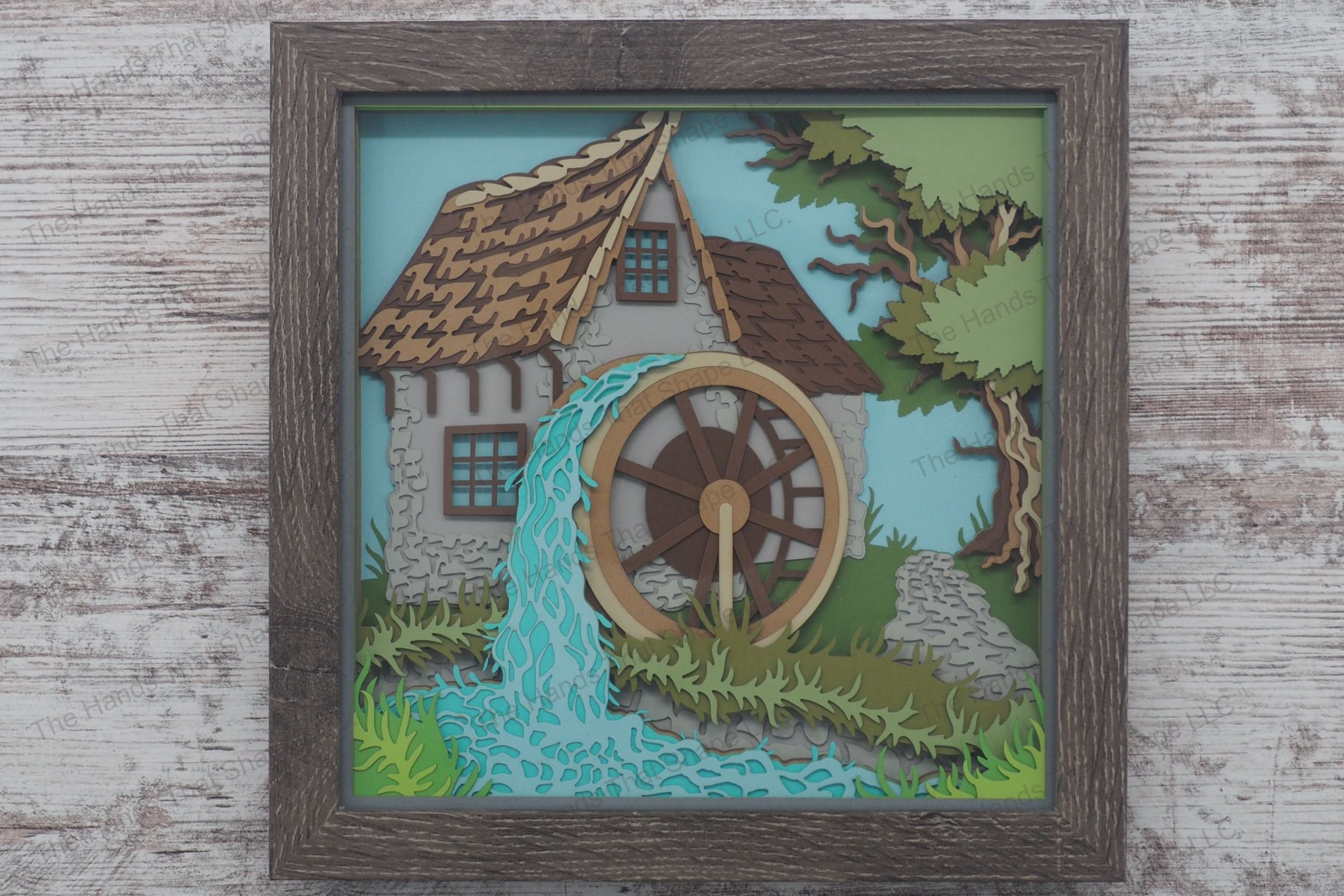 A framed layered paper shadowbox depicting a rustic water mill with a shake roof, stone walls, and water flowing over the wheel by a stream.