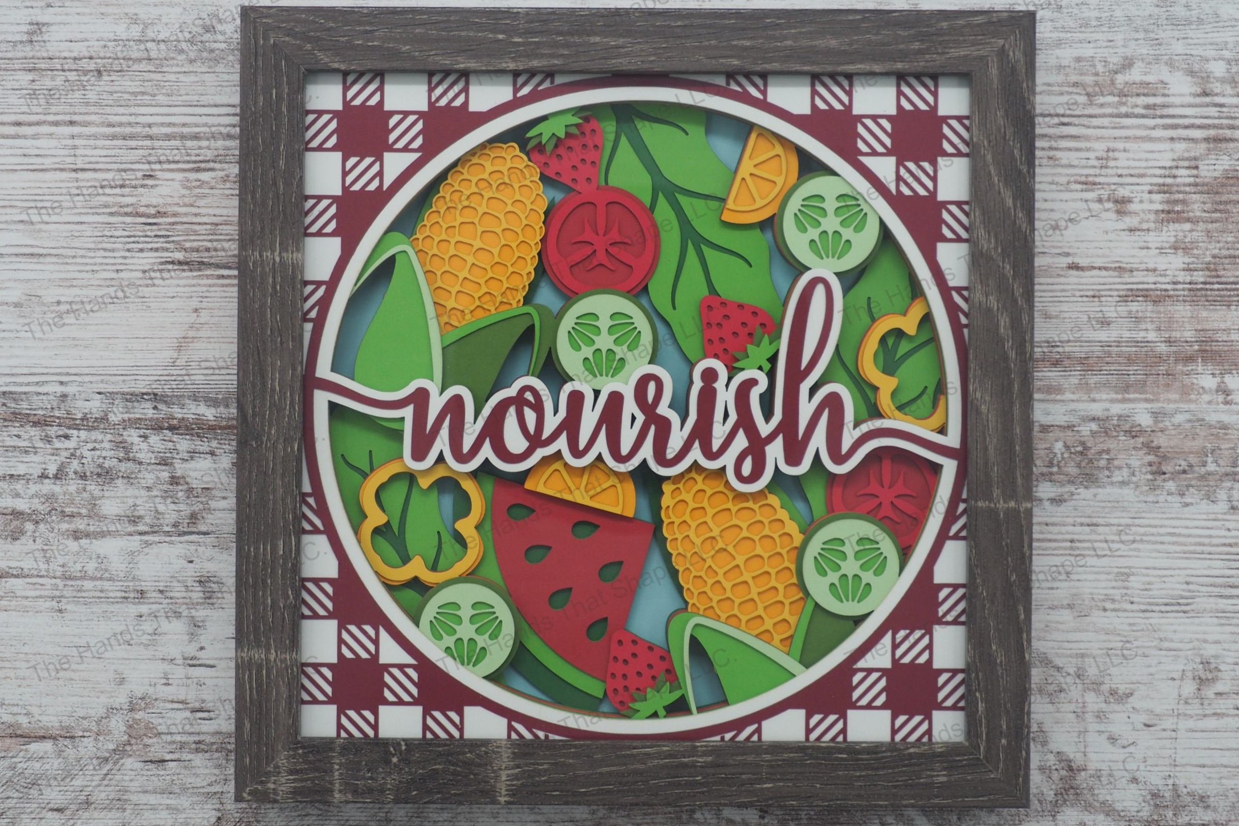 A vibrant layered paper shadowbox depicting a harvest scene with fruits and vegetables arrayed around the word 'nourish', framed by a picnic tablecloth pattern.
