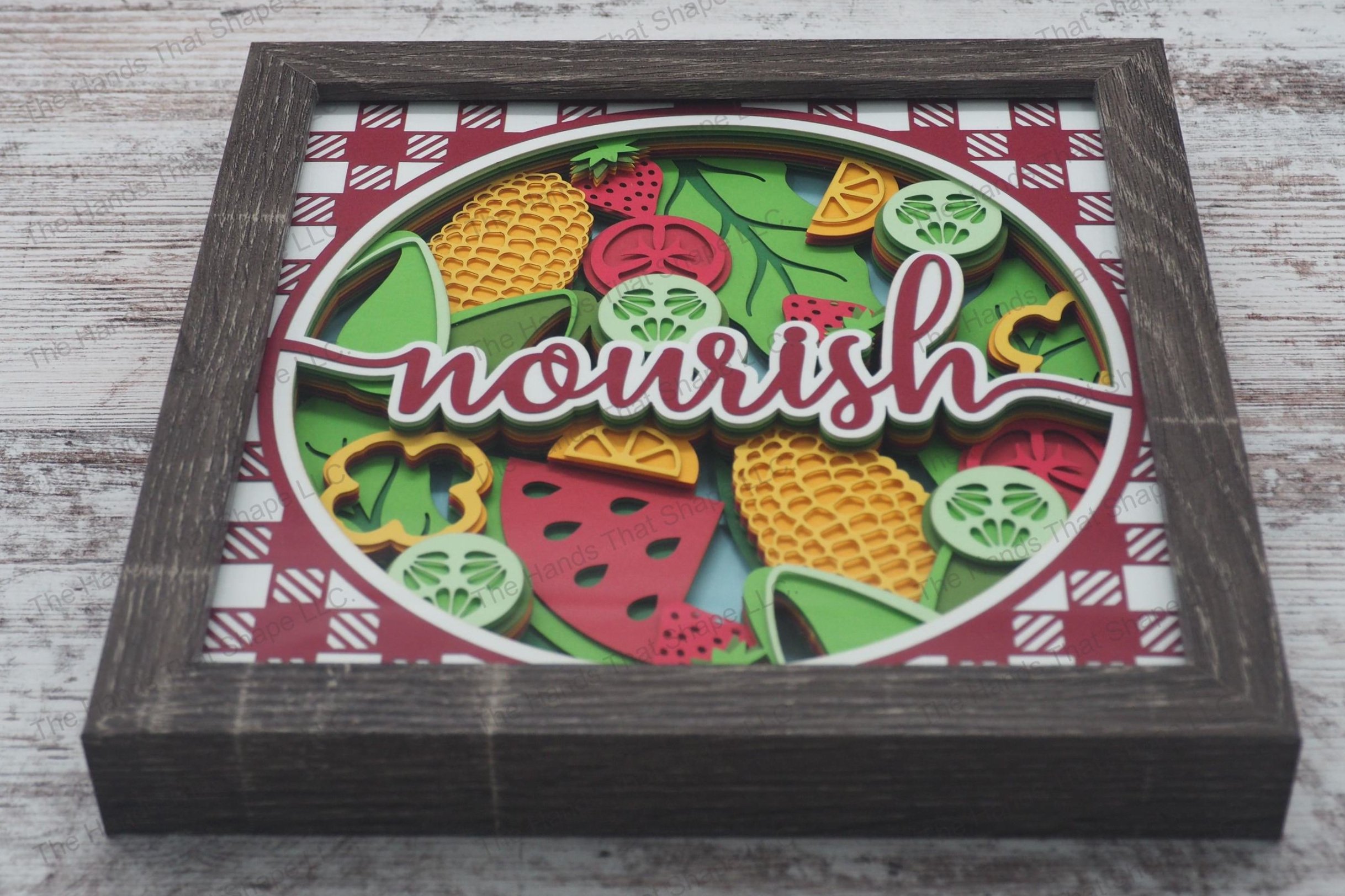 A vibrant layered paper shadowbox depicting a harvest scene with fruits and vegetables arrayed around the word 'nourish', framed by a picnic tablecloth pattern.