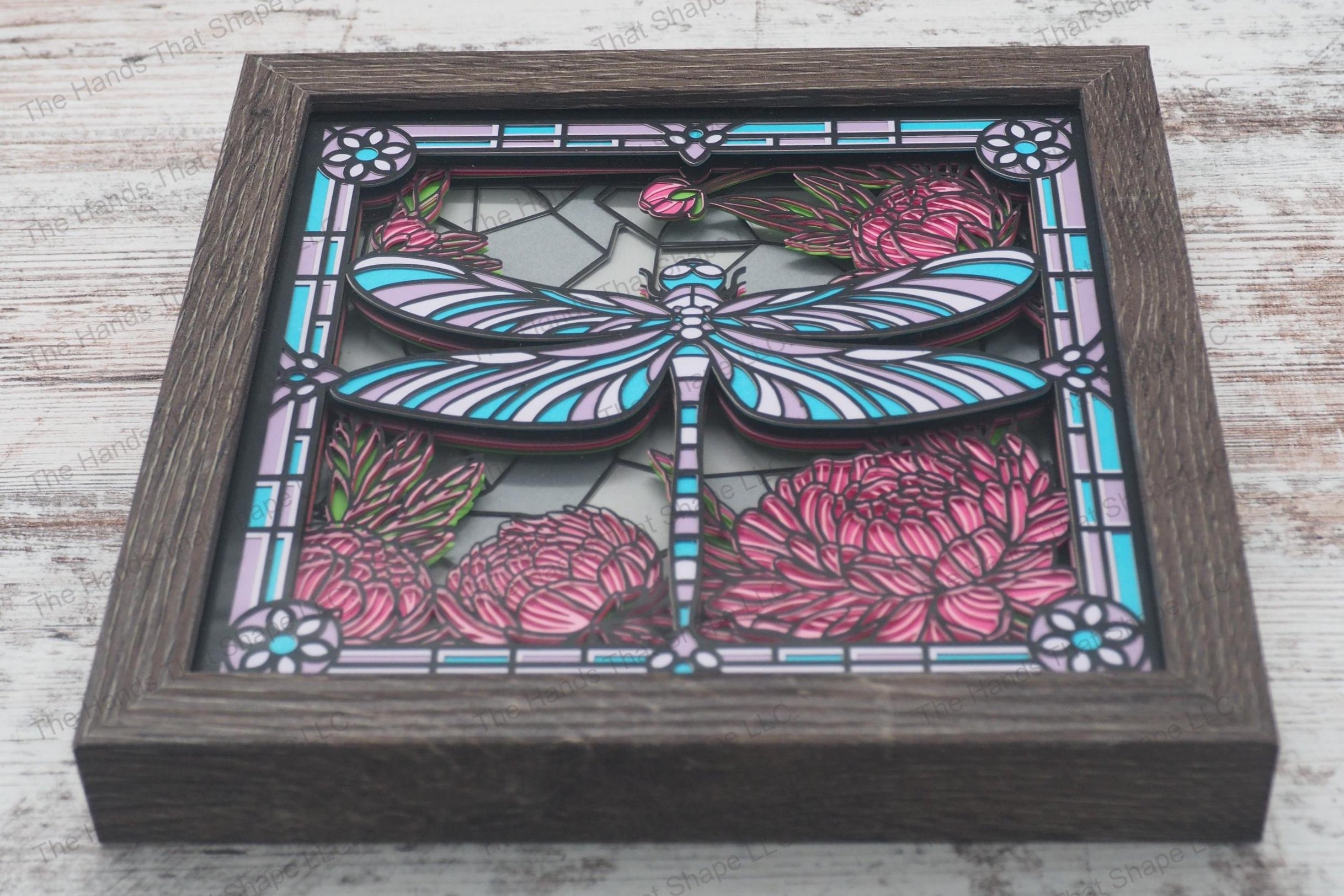 A framed layered paper shadowbox depicting a dragonfly hovering over a blooming garden, crafted in a stained glass style, made from 11 layers of cardstock.