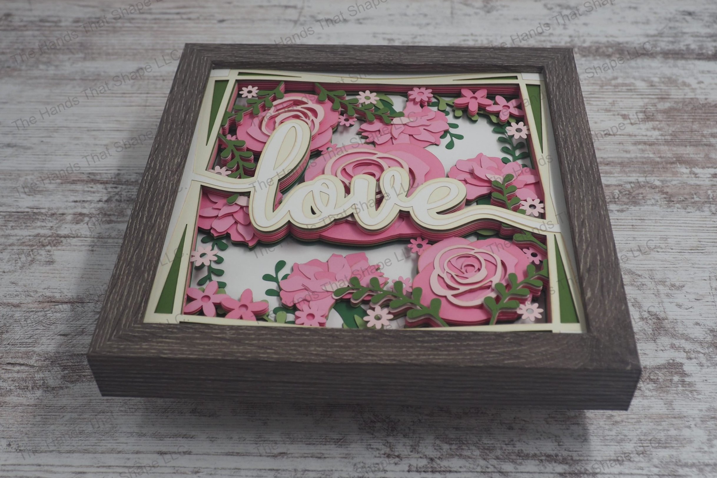 A romantic layered paper shadowbox featuring the word 'Love' surrounded by vibrant, multi-layered roses and greenery, crafted from cardstock in a gray frame.
