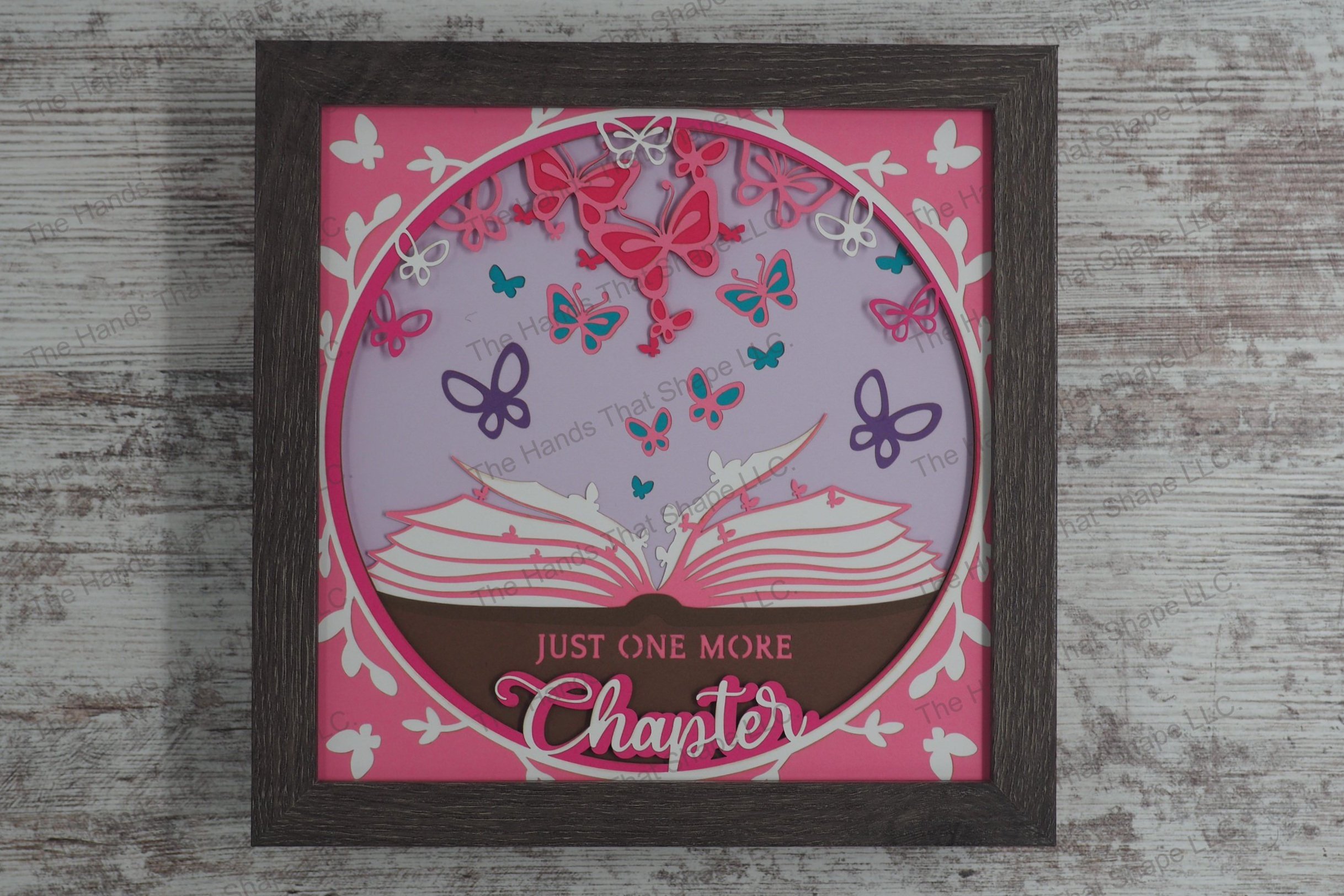 A layered paper shadowbox depicting an open book with colorful butterflies emerging from the pages, accompanied by the phrase 'Just one more chapter.'