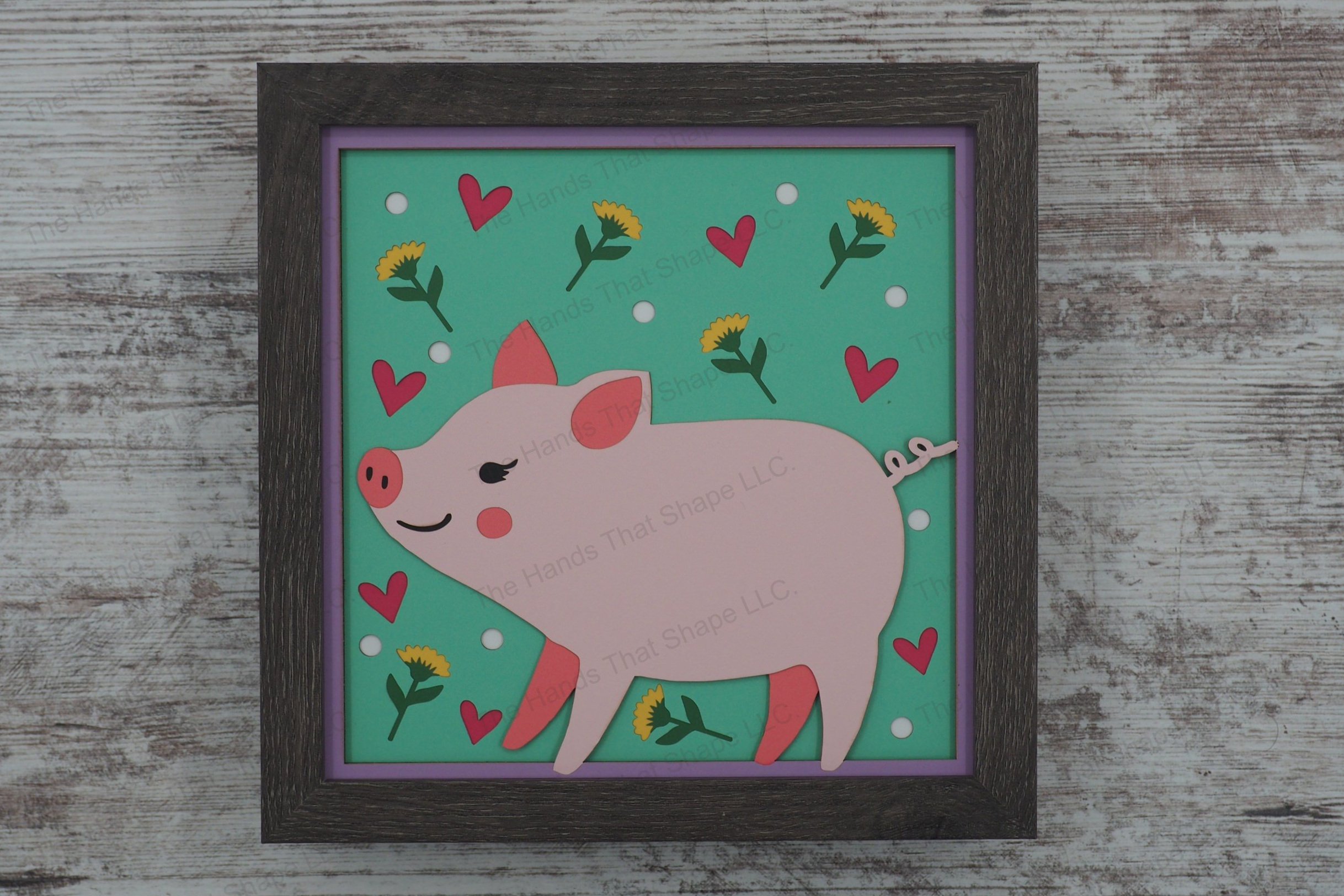 This layered paper shadowbox features a folksy piglet, surrounded by charming flowers, dots, and hearts, perfect for adding whimsical charm to any room.