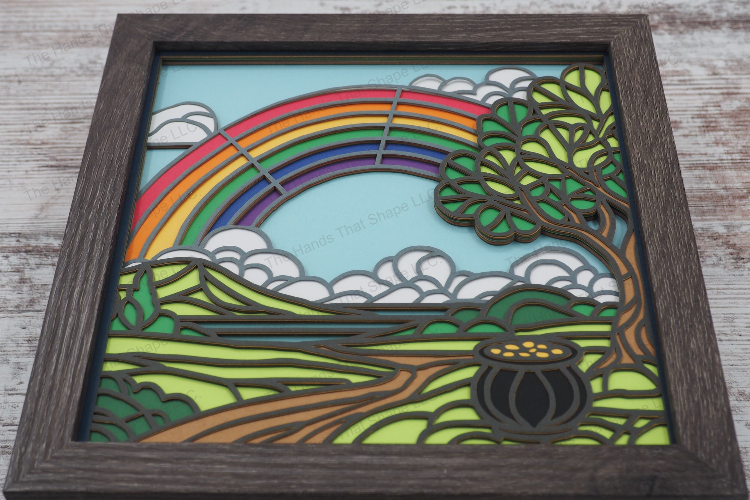 A vibrant layered paper shadowbox featuring a pot of gold, a winding path leading to a distant lake, and a brilliant rainbow arching across the sky.