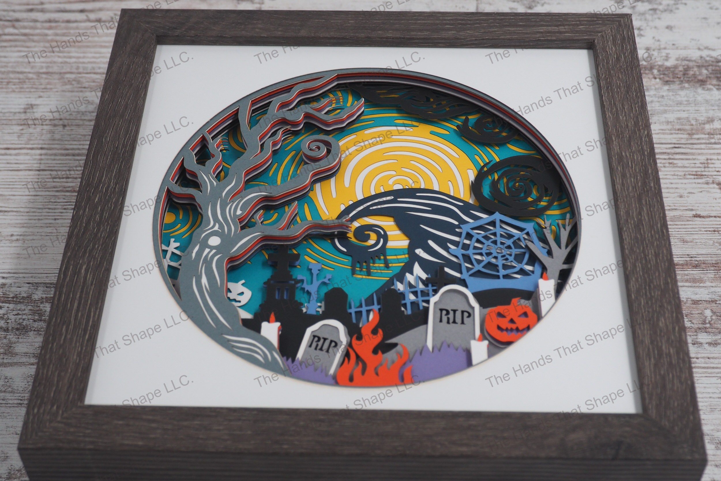 A captivating layered paper framed shadowbox depicting a spooky Halloween cemetery scene with aged tombstones, an ancient tree and a witch's hat-shaped hill.