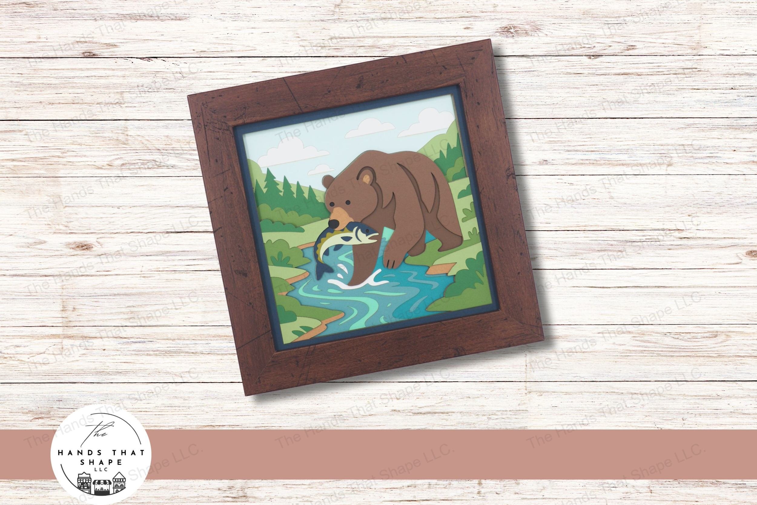 This layered cardstock art piece features a powerful grizzly bear standing in a stream with a fish in its mouth, set against a lush forest backdrop.