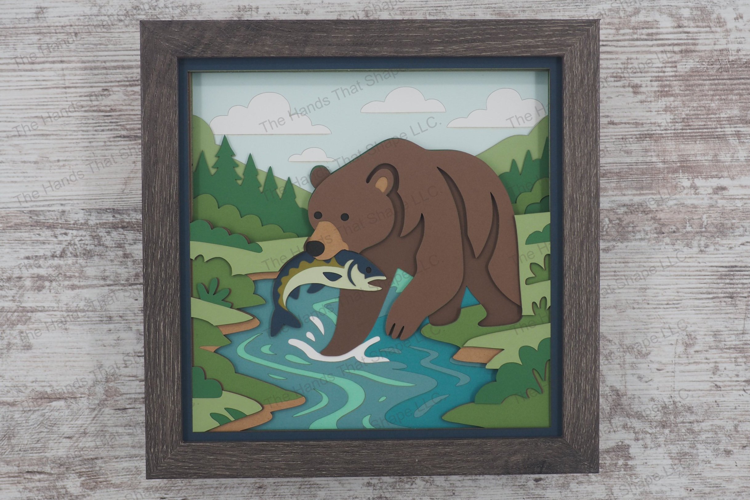 A dramatic 3D layered paper shadowbox featuring a grizzly bear standing in a stream with a fish in its mouth, framed by lush greenery, trees, and a cloudy sky.