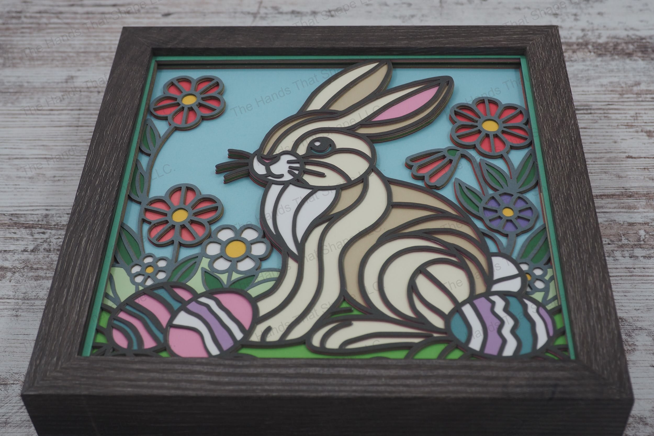 A charming framed layered paper shadowbox featuring a bunny sitting in a grassy meadow surrounded by colorful Easter eggs and blooming spring flowers.