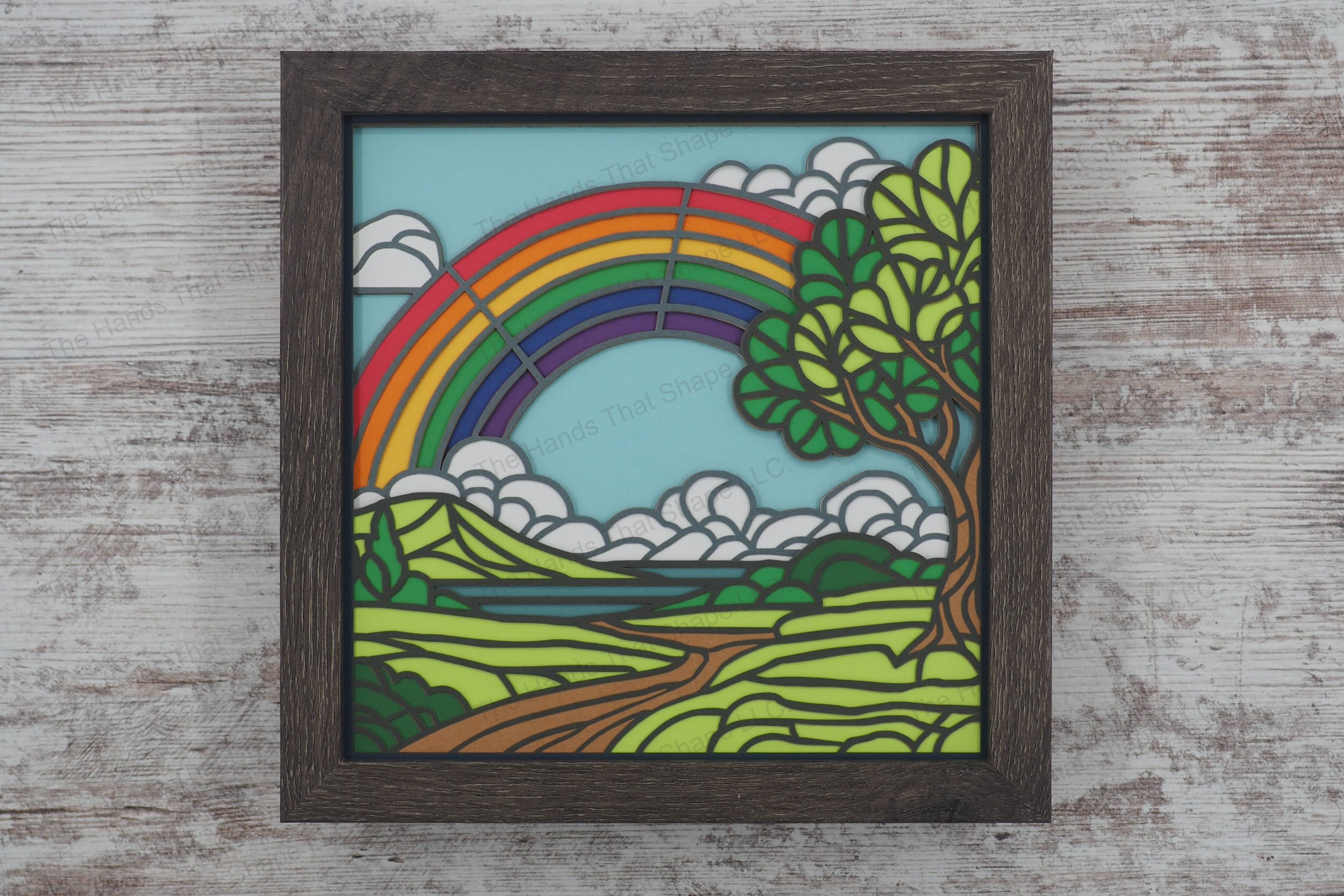 This layered paper shadowbox features a vibrant rainbow arching over a picturesque countryside with rolling hills, and a winding path to a distant lake.