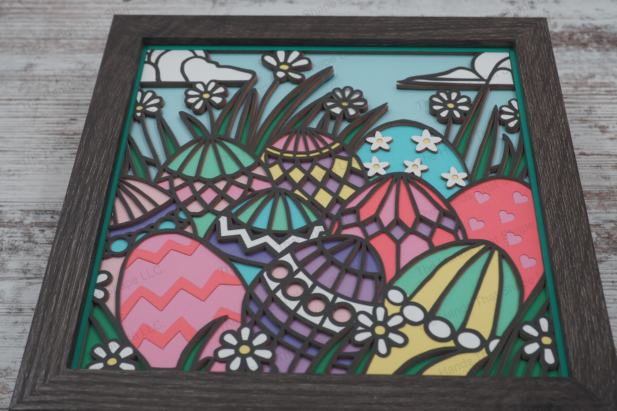 A vibrant layered paper shadowbox featuring intricately decorated Easter eggs nestled in lush grass, surrounded by blooming daisies and a cloud-filled sky.
