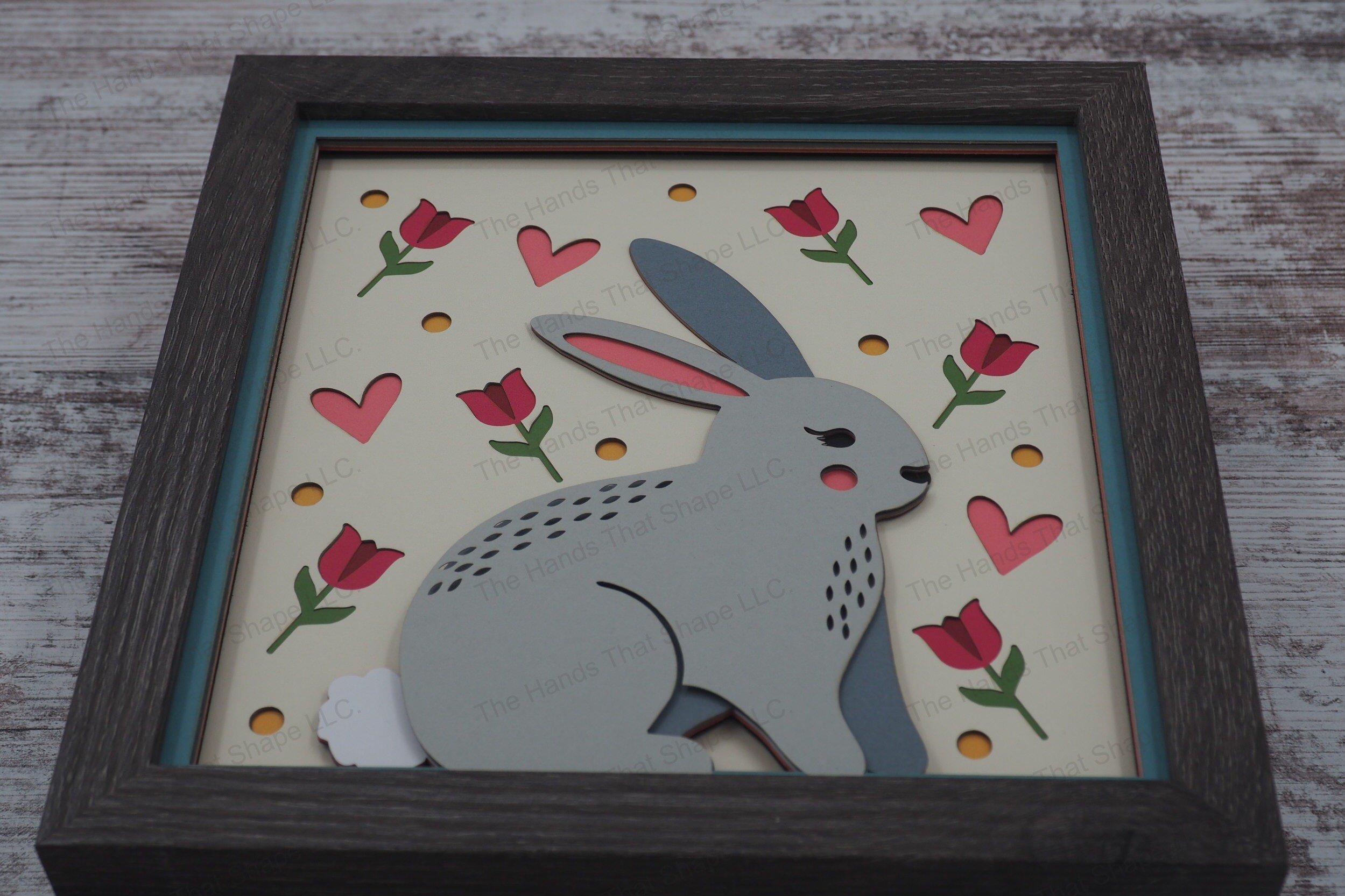 Charming layered paper shadowbox features a folksy bunny surrounded by tulips, hearts, and dots, perfect for bringing springtime warmth and joy to any space.
