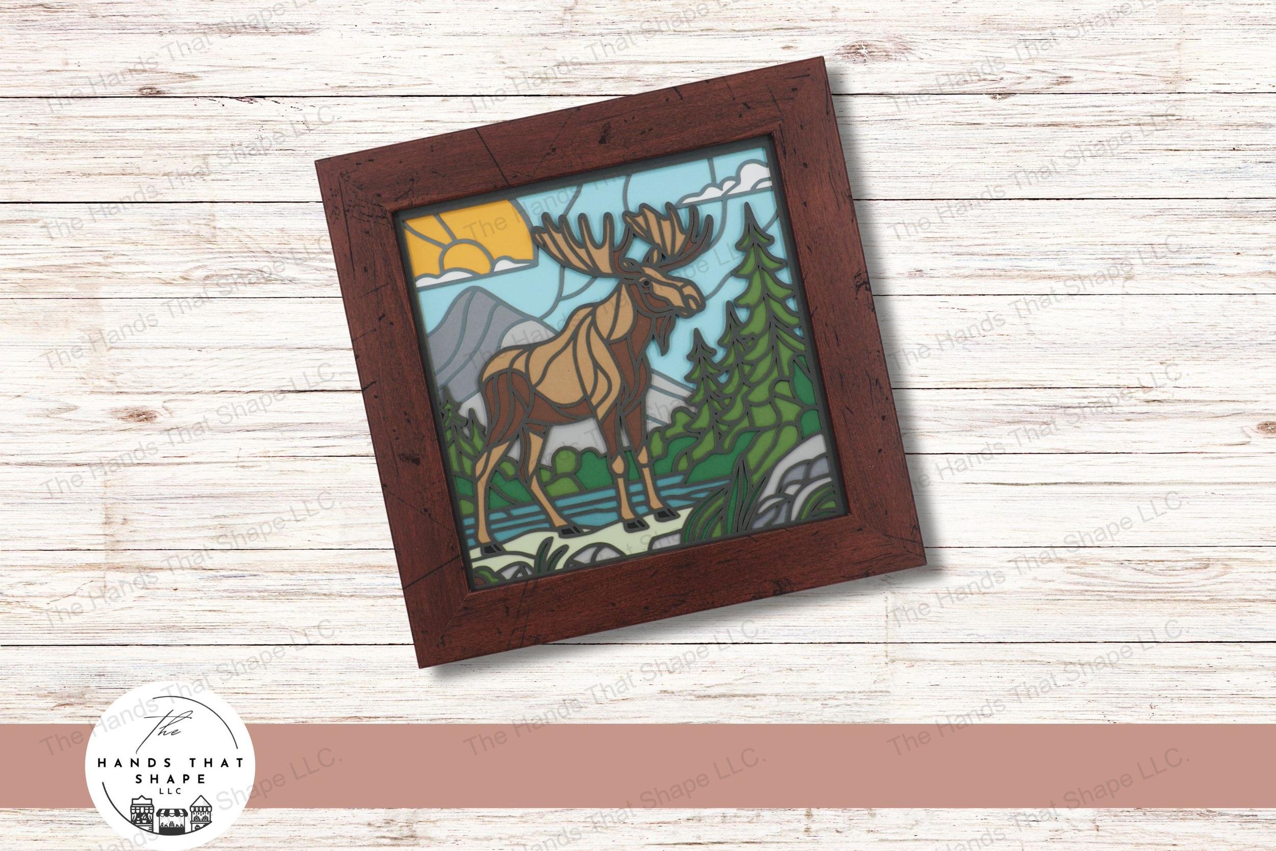 This layered cardstock art piece vividly captures a moose by the water’s edge, surrounded by mountains and trees, creating a striking stained glass effect.