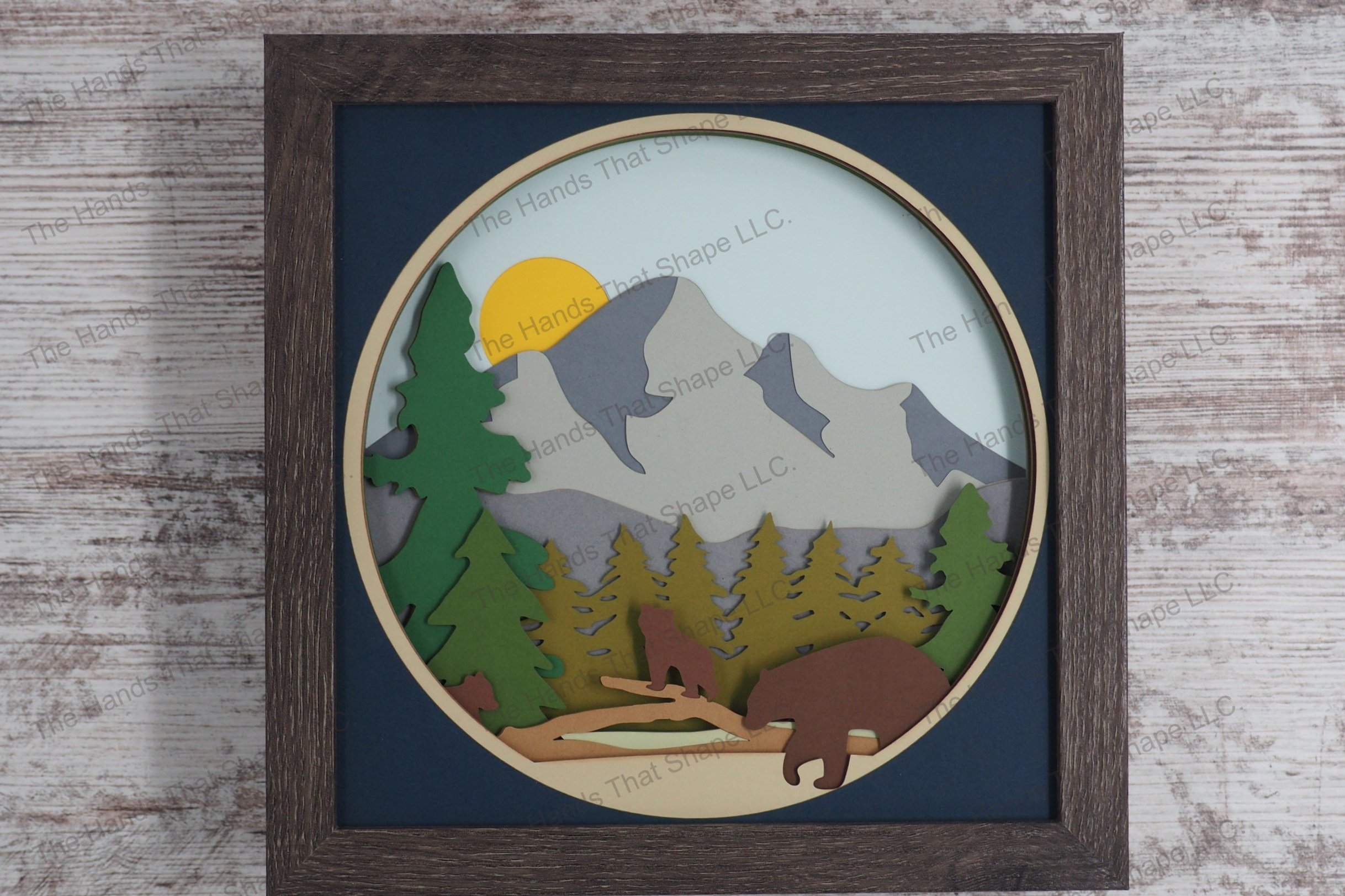 A charming depiction of a bear family interacting in a mountain forest, this layered paper shadowbox captures mama bear and her cubs among pine trees.