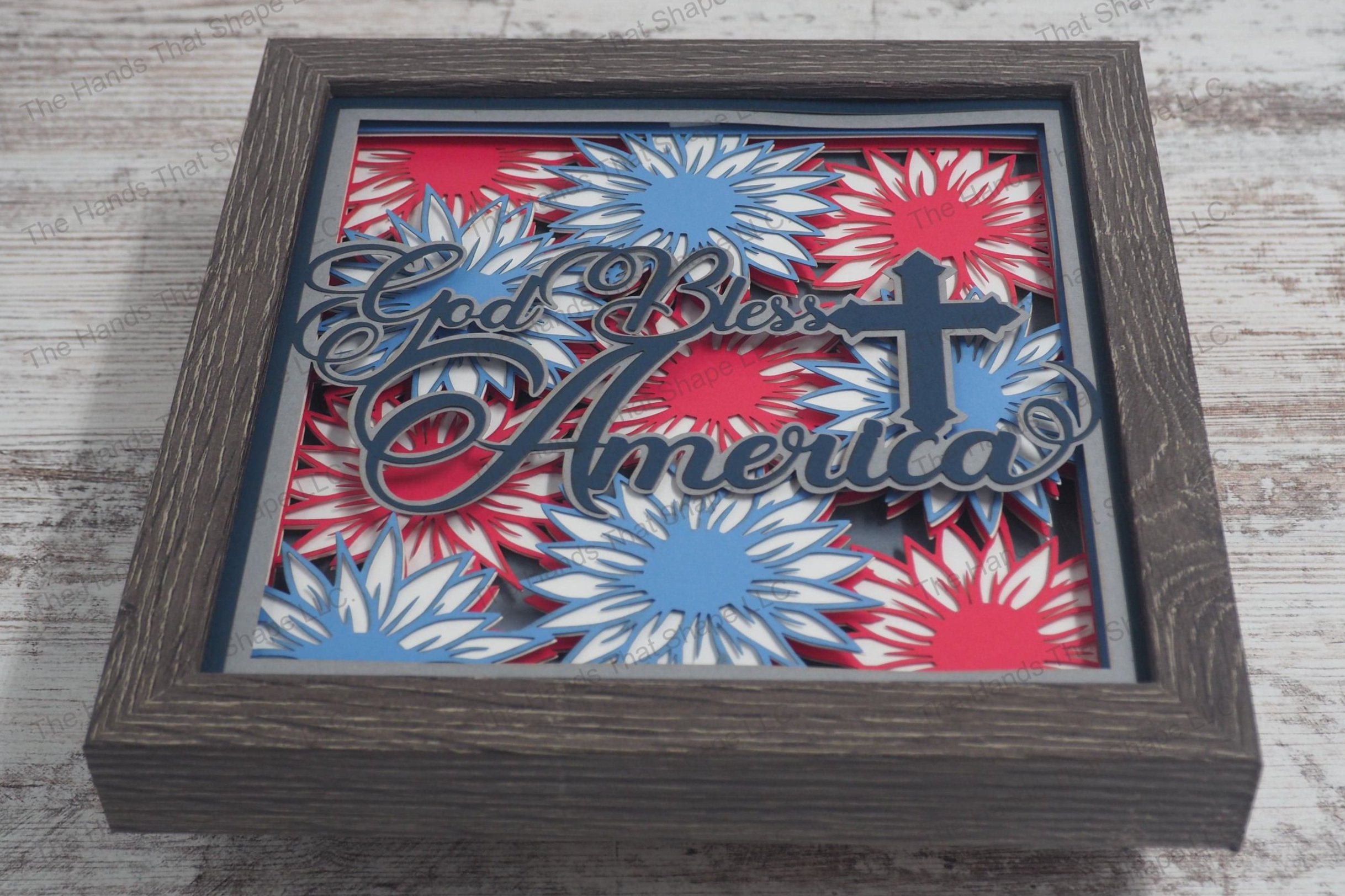 A layered paper shadowbox featuring a patriotic display of blue, white, and red flowers resembling fireworks, available with the text 'God Bless America'.