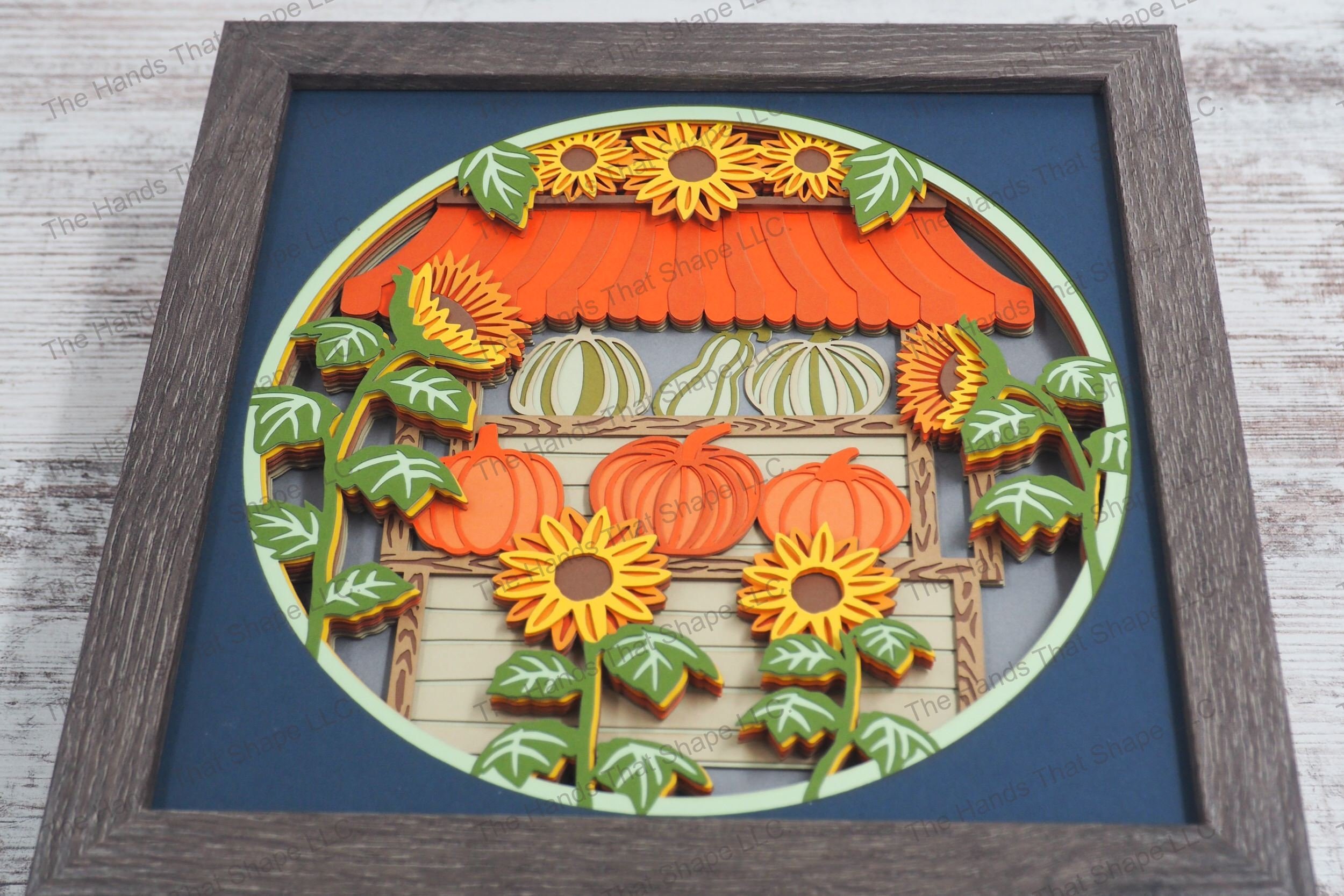 A vibrant framed layered paper shadowbox depicting a market stand filled with pumpkins and surrounded by sunflowers, crafted from 15 layers of cardstock.