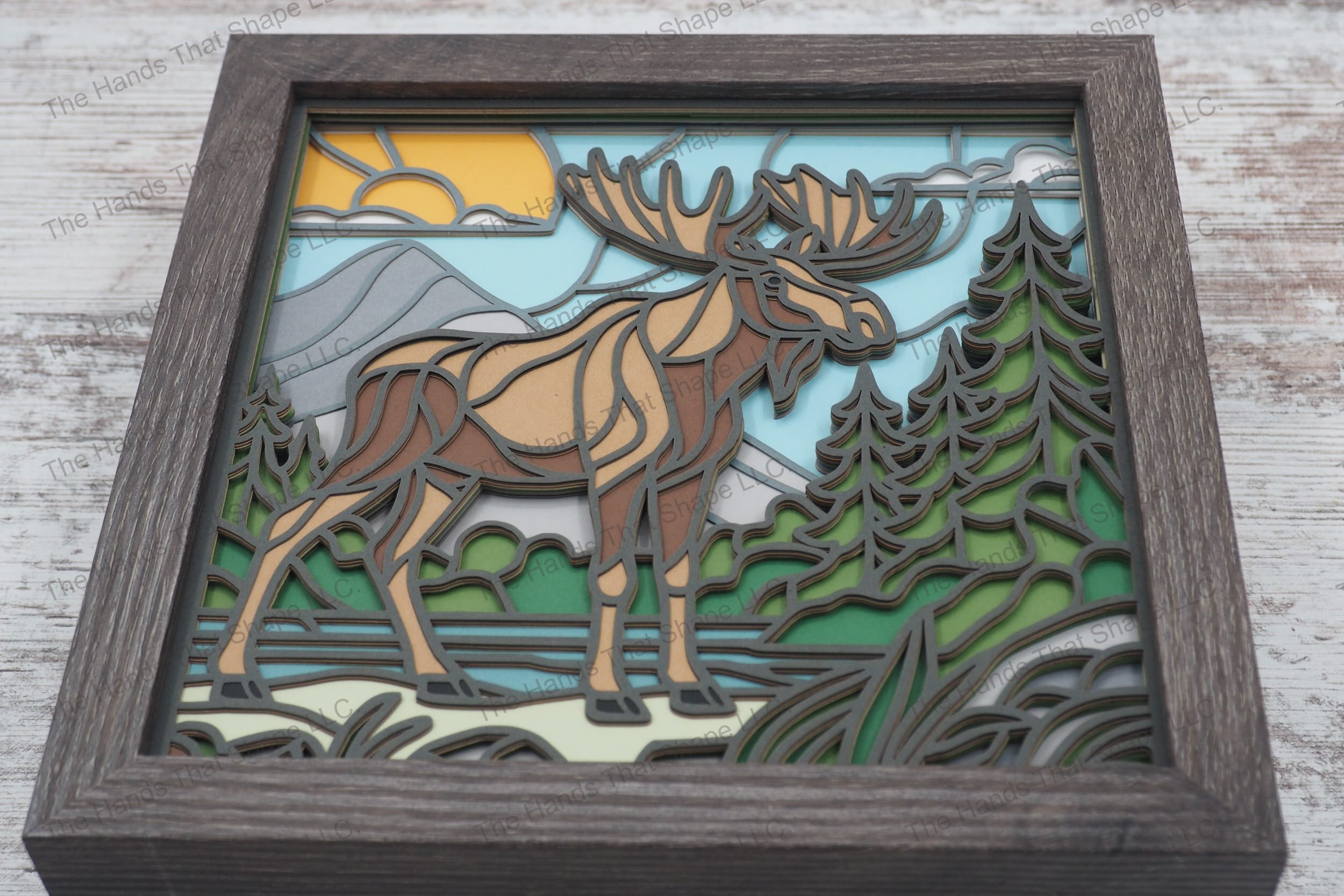 Explore the beauty of the wilderness with this layered paper shadowbox, featuring a moose at a water's edge, set against a backdrop of pine trees and mountains.