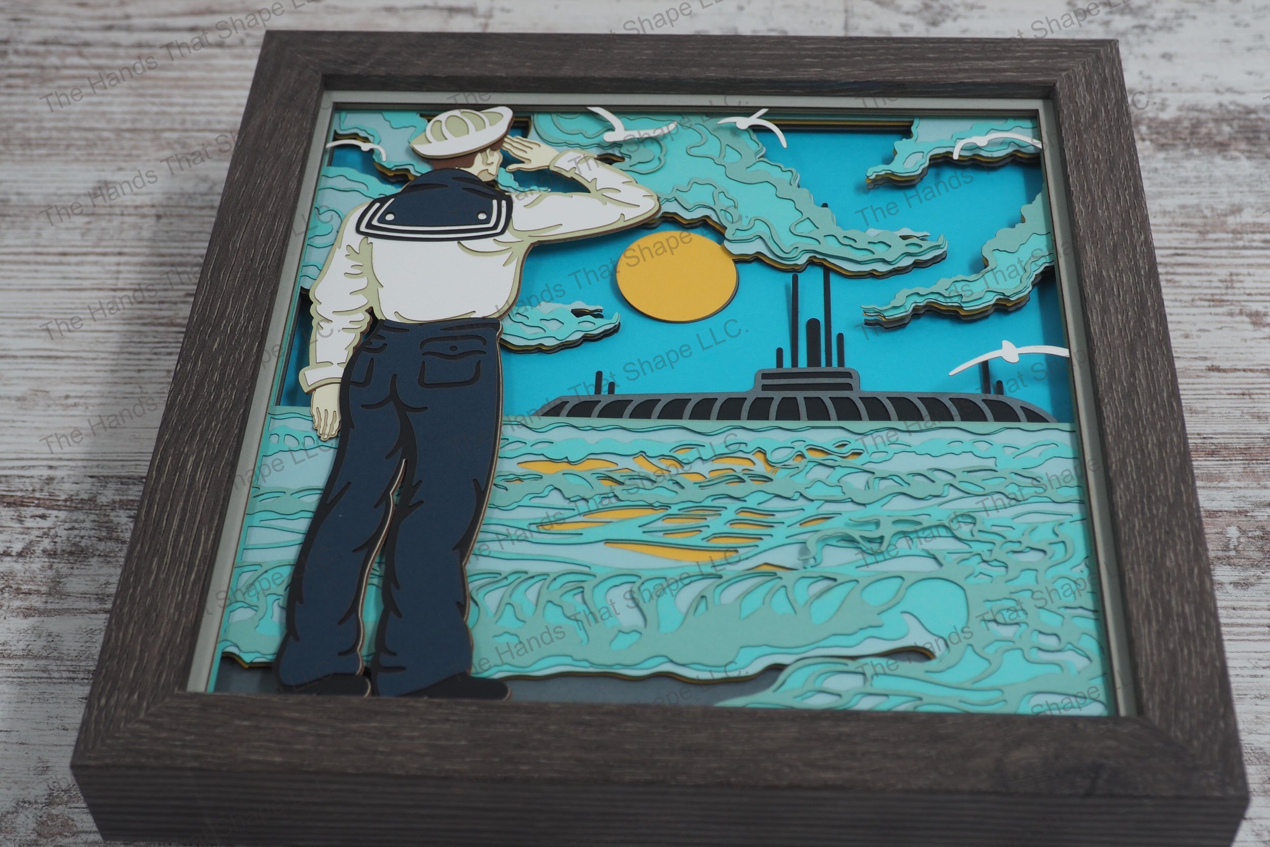 A framed layered paper shadowbox depicting a saluting sailor and a floating submarine under a radiant sun, with seagulls soaring above.