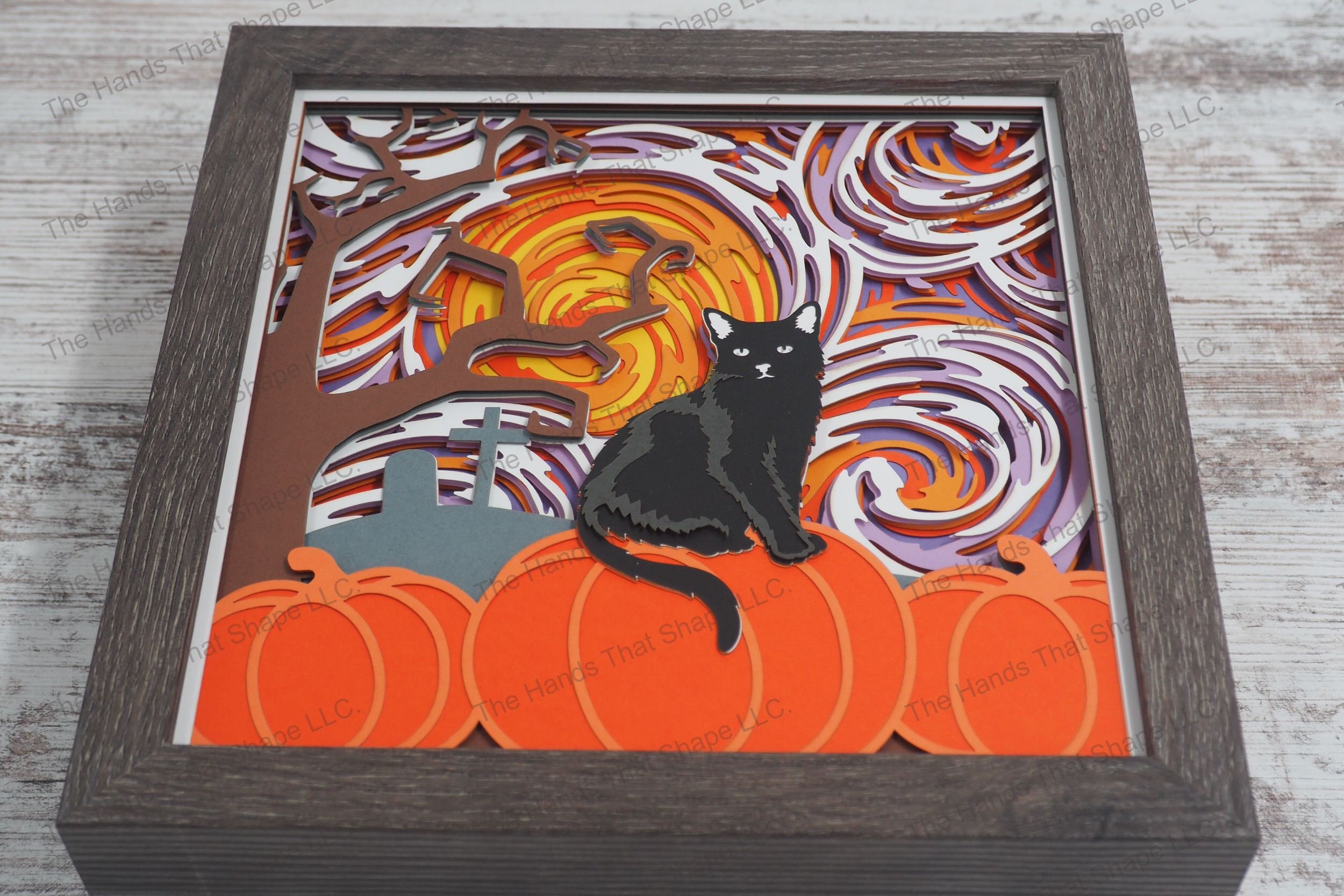 A framed layered paper shadowbox depicting a Halloween scene with a black cat perched on a pumpkin under a swirling, colorful sky on a full moon night.