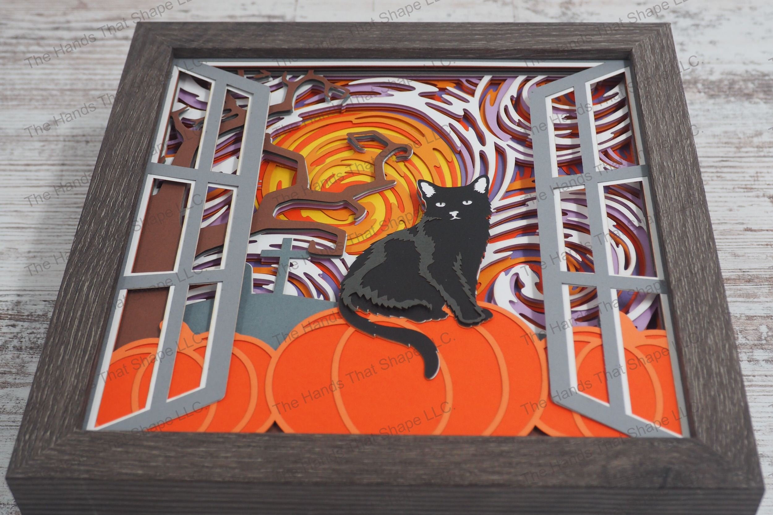 A layered paper shadowbox depicting a Halloween scene through a window with a black cat perched on a pumpkin under a swirling, colorful sky on a full moon night