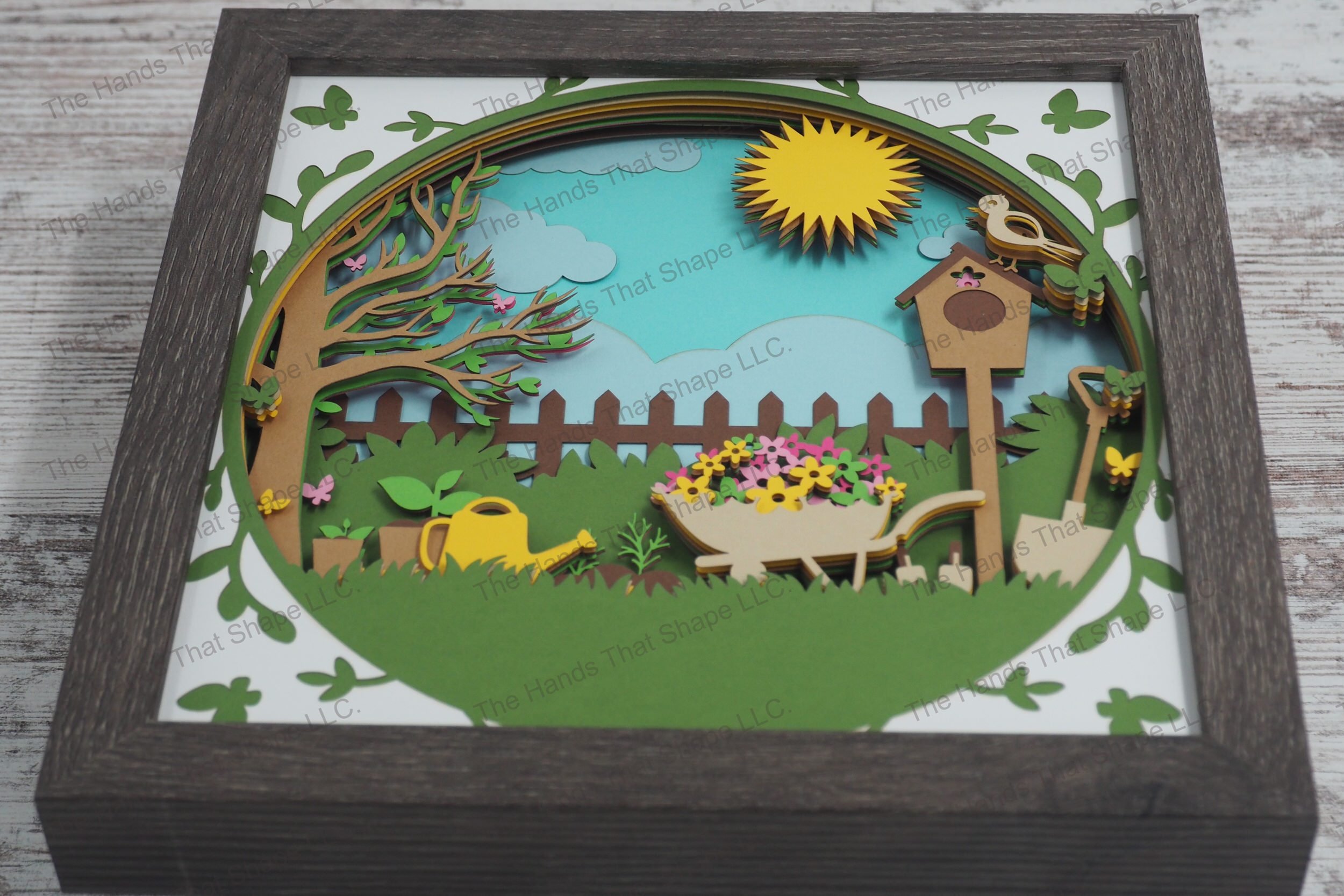 A detailed layered paper shadowbox depicting a lush backyard garden scene with a wheelbarrow full of flowers, gardening tools, butterflies, and a sunlit sky.