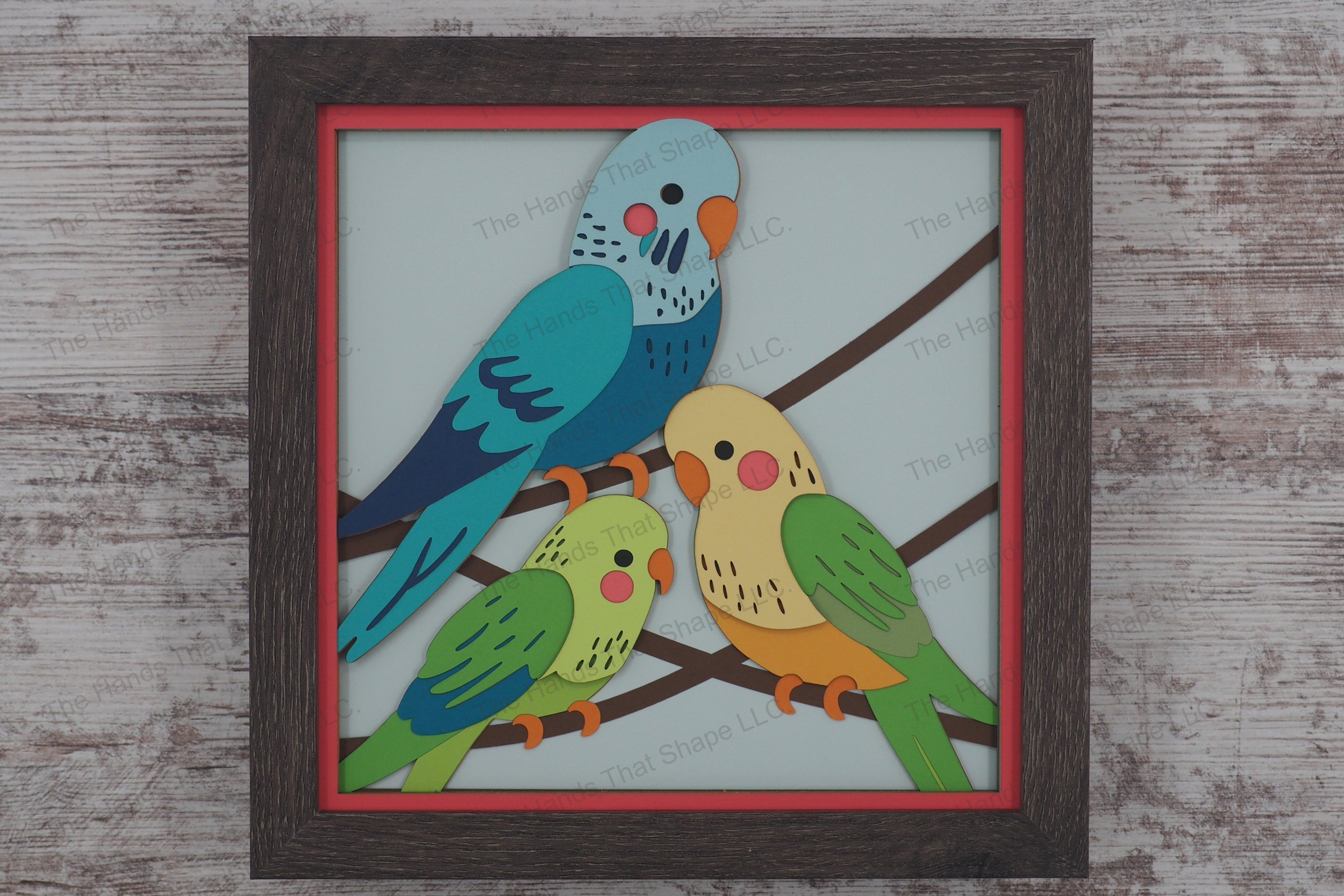A colorful 3D layered paper shadowbox featuring three uniquely feathered parakeets perched on vines, brought to life with 20 vibrant cardstock layers.