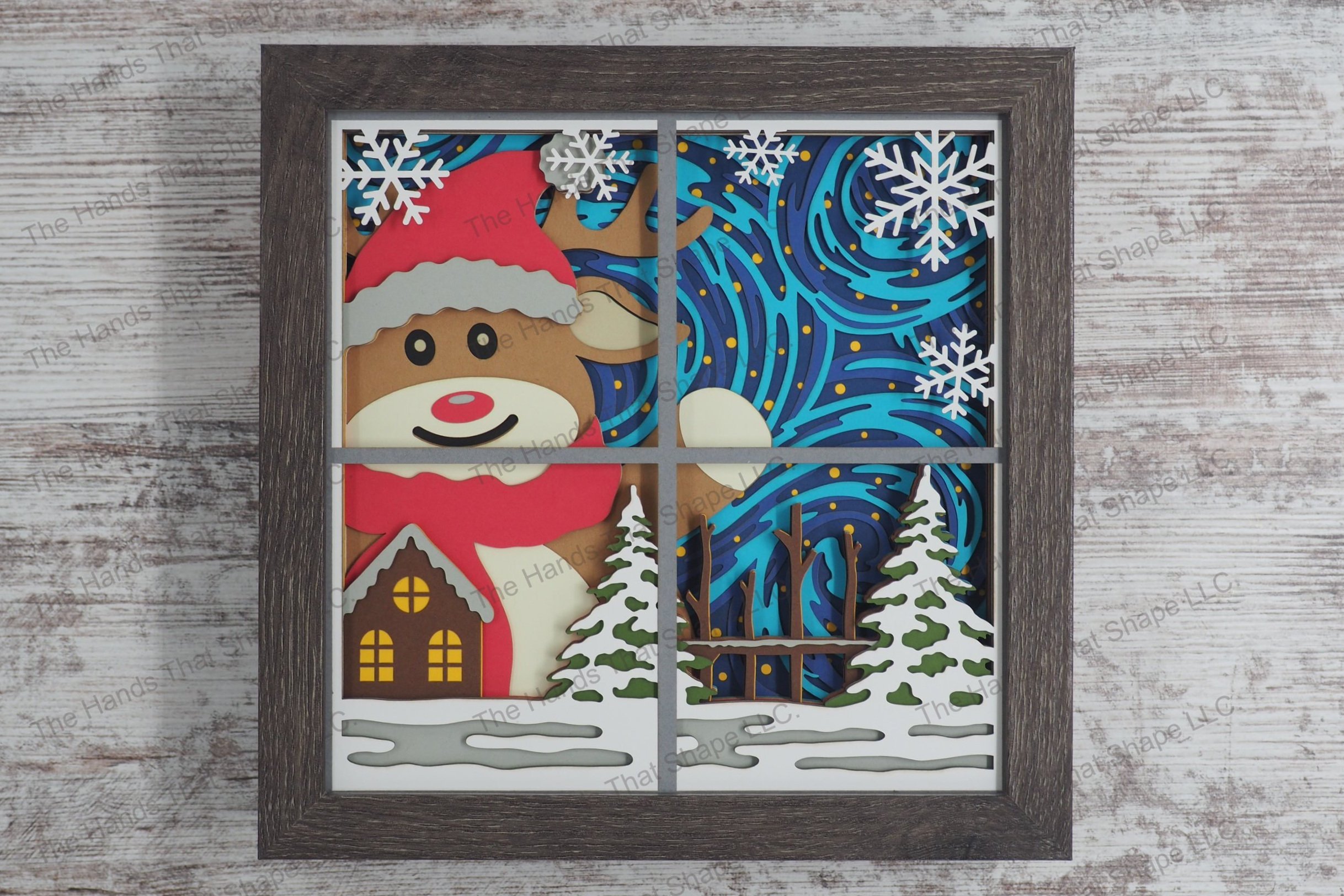 A layered paper shadowbox depicting an oversized festive reindeer through a window waving joyfully, with wintery swirls and stars as a backdrop.